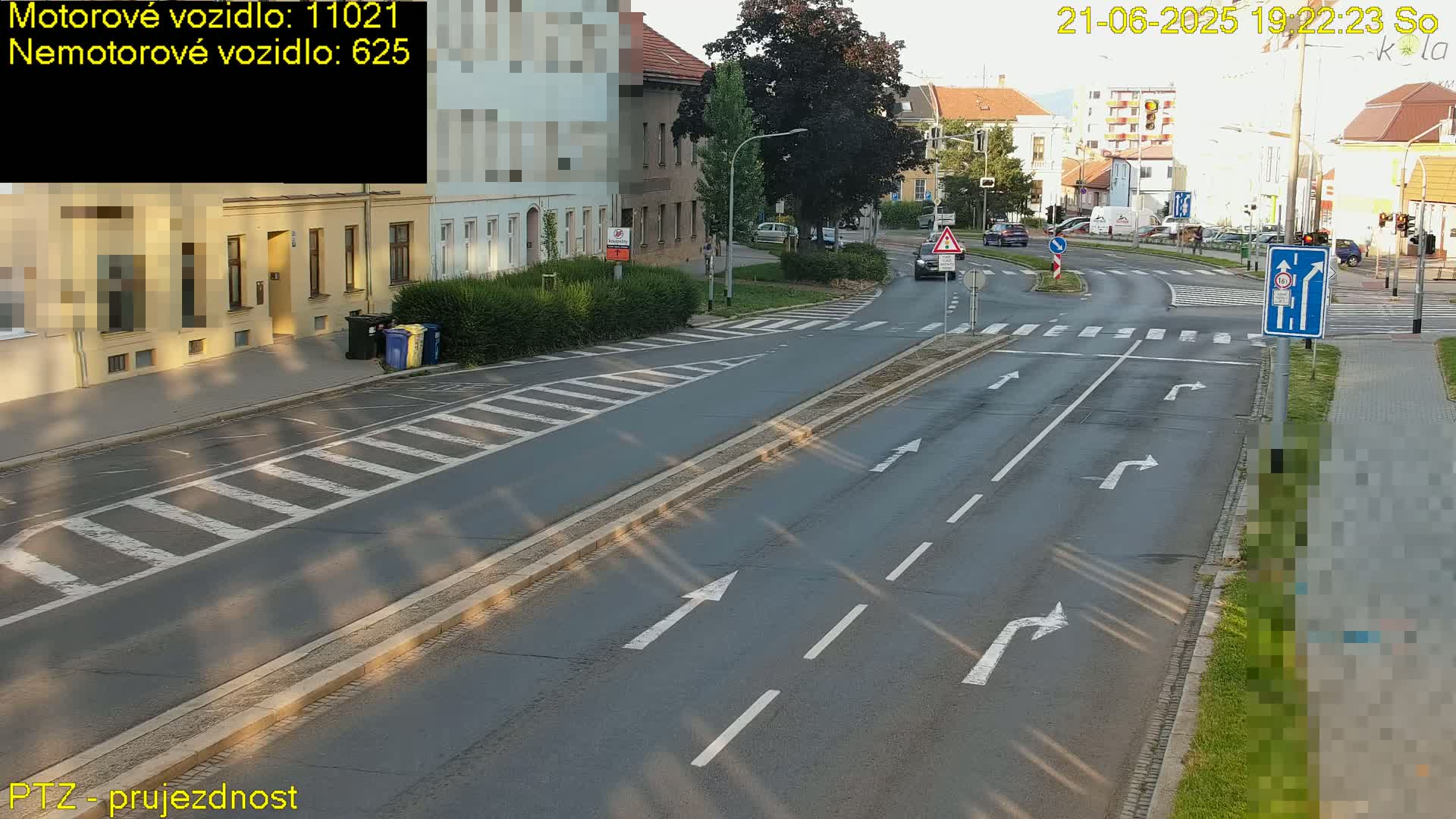 A sunny street scene shows a multi-lane road with crosswalks and turning lanes, several buildings and parked cars visible in the background.