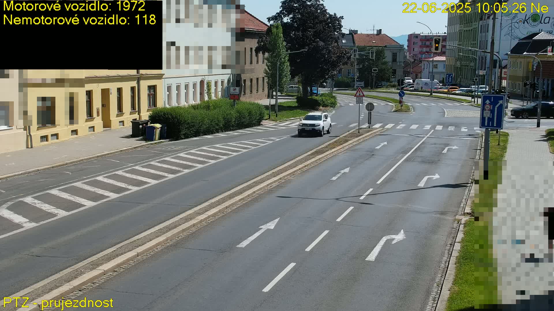 A sunny day shows a multi-lane road with a white SUV driving, pedestrian crossings, and buildings lining the street.