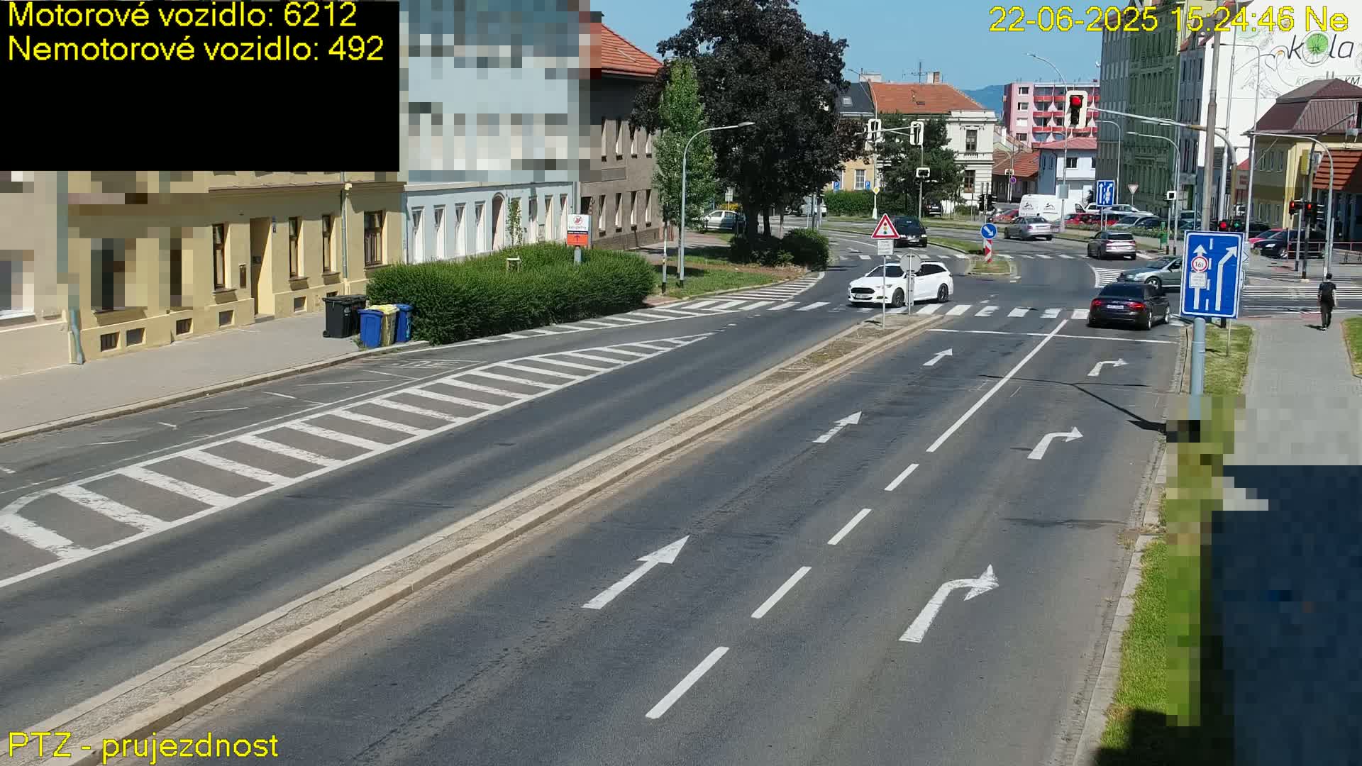 A sunny street scene shows several cars at an intersection with crosswalks and lane markings, along with buildings and some pedestrians.