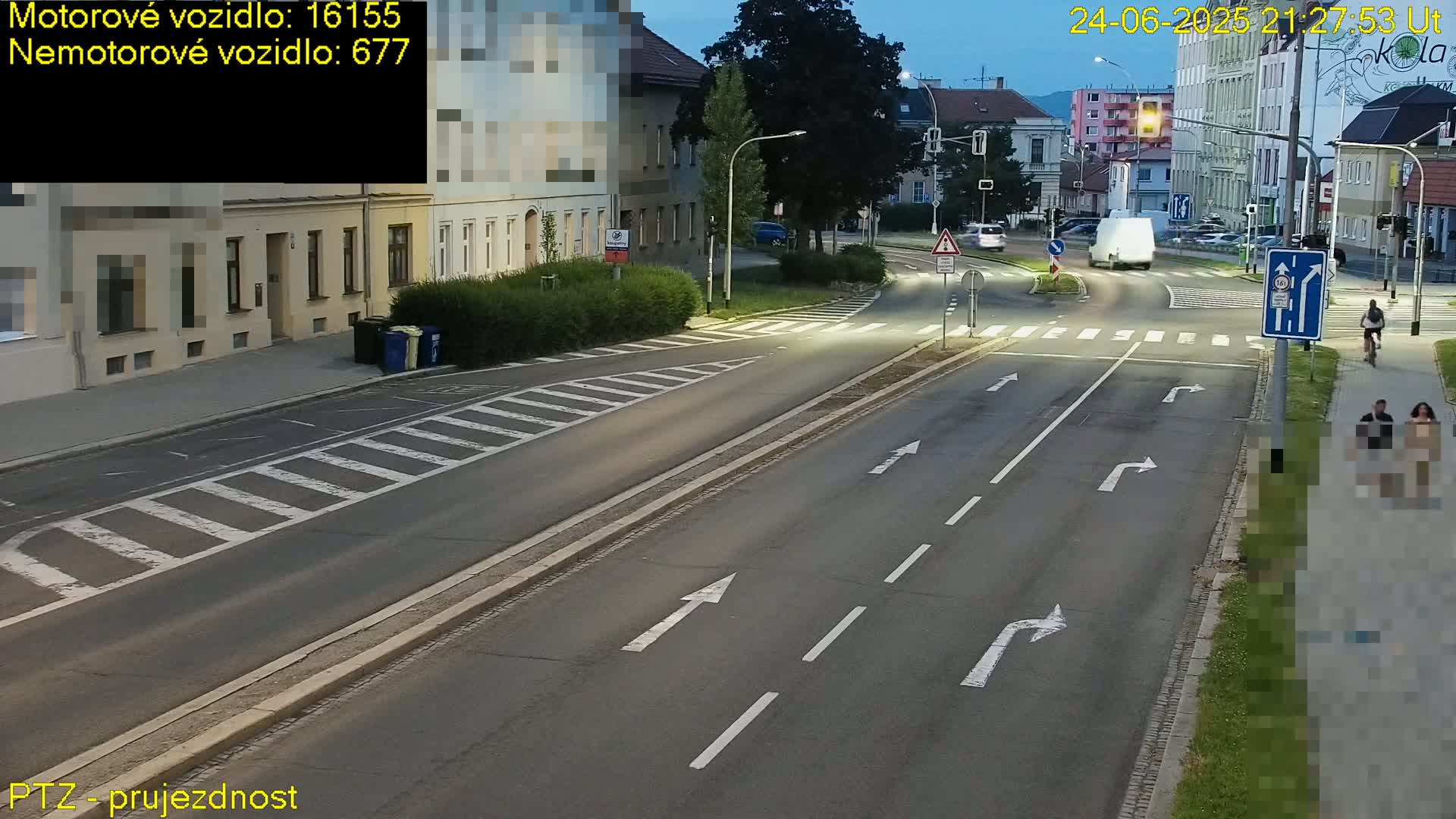 A mostly empty asphalt road with pedestrian crosswalks intersects with another road at dusk, with a few vehicles and pedestrians visible.
