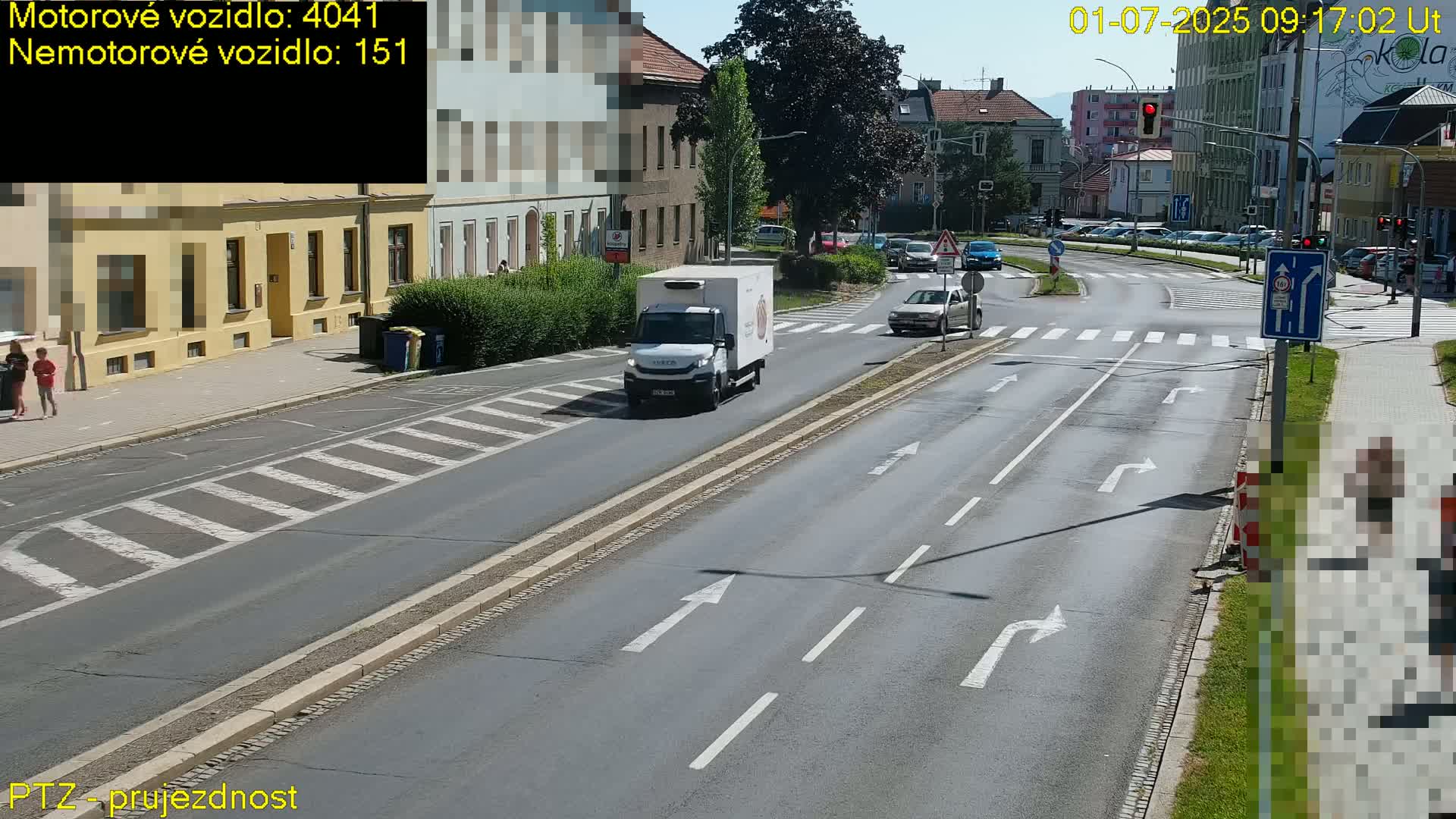 A sunny day shows a white delivery truck driving on a multi-lane road that intersects with another road, several pedestrians are visible on the sidewalks, and cars are parked along the side of the road.