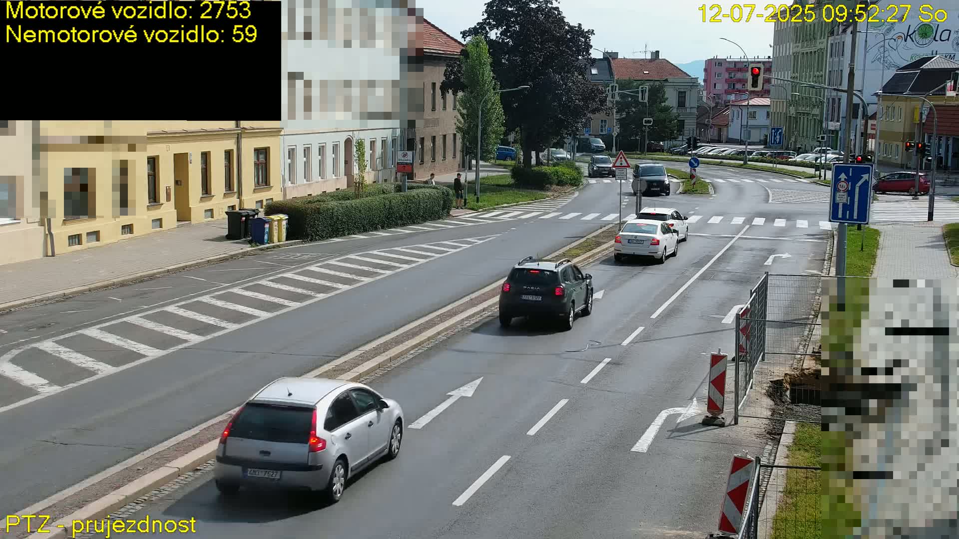 A sunny day shows a white delivery truck driving on a multi-lane road that intersects with another road, several pedestrians are visible on the sidewalks, and cars are parked along the side of the road.