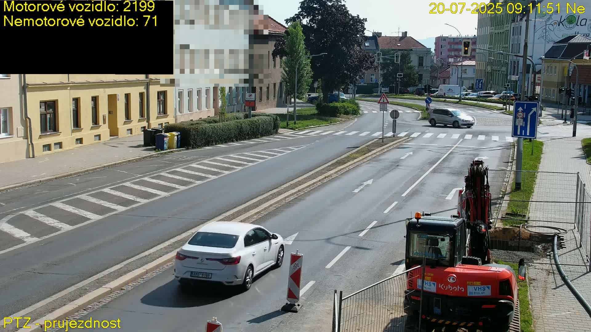 A sunny day on a city street shows a white car driving past a small orange construction vehicle near a crosswalk.