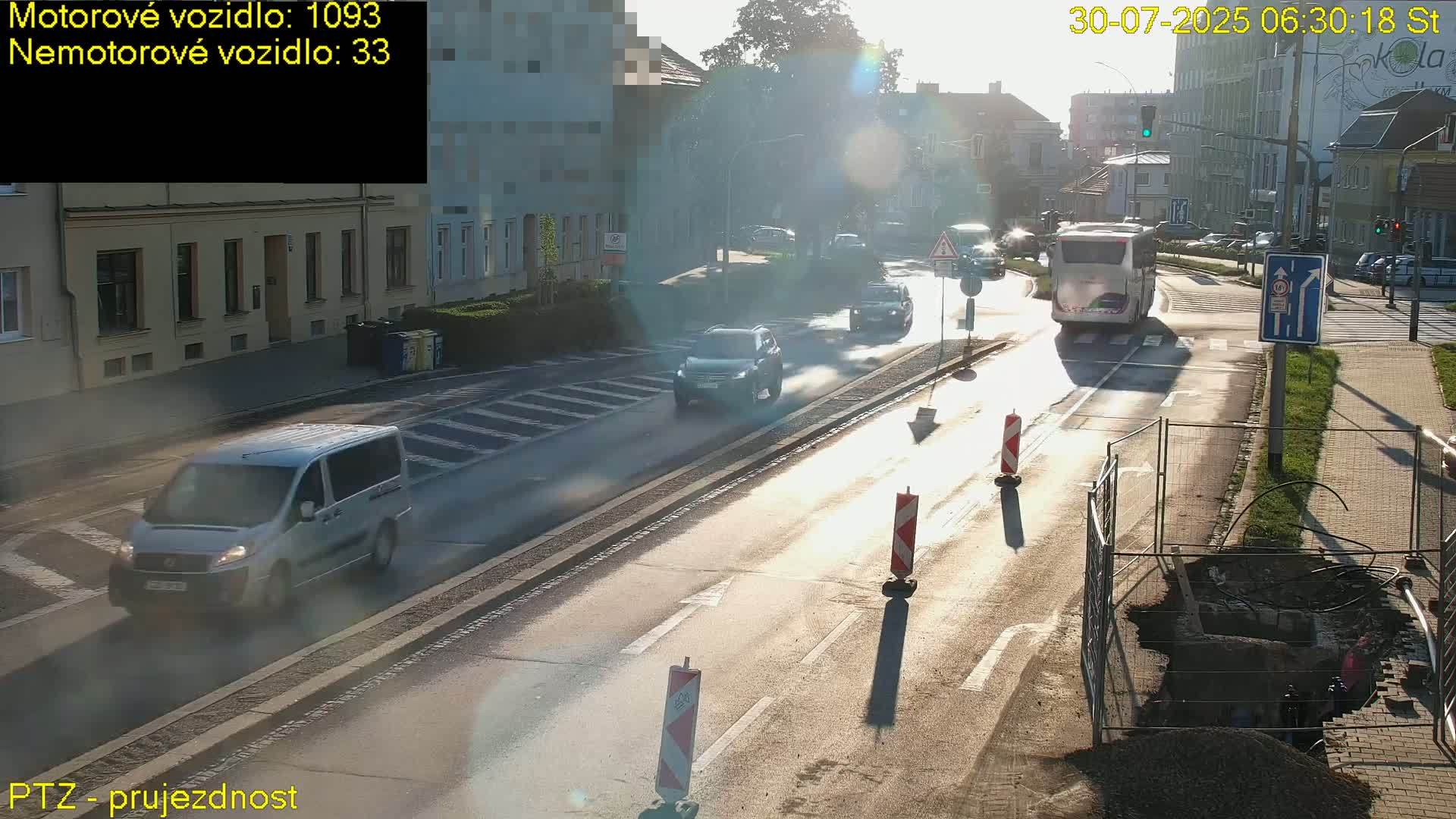 A sunny day on a city street shows a white car driving past a small orange construction vehicle near a crosswalk.