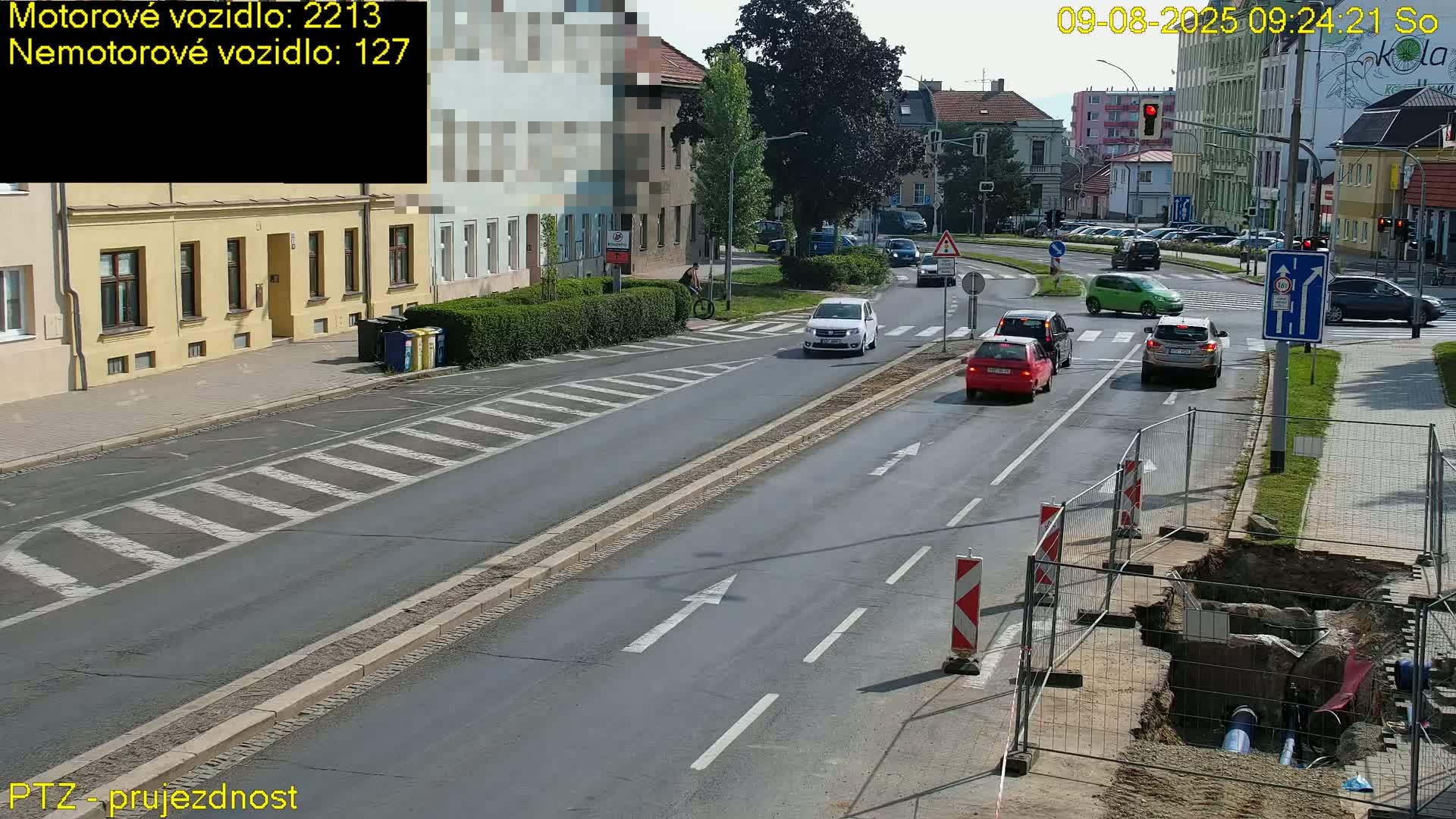 A sunny day shows several cars driving on a road with a crosswalk and roadwork visible on the side, including a dug-up section fenced off.