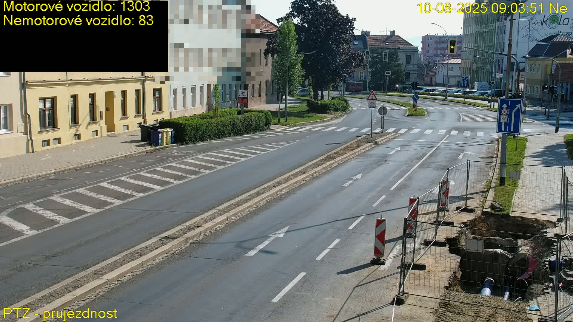 A sunny day shows a street scene with a road under construction, featuring a dug-up section cordoned off by a temporary fence.