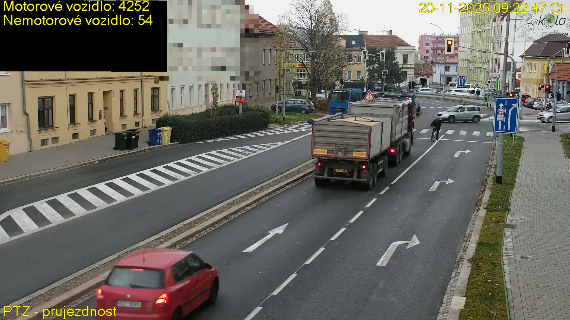 On a cloudy day, a dump truck and a red car drive down a multi-lane city street towards an intersection where a cyclist crosses, with several other vehicles and buildings visible under overcast skies.
