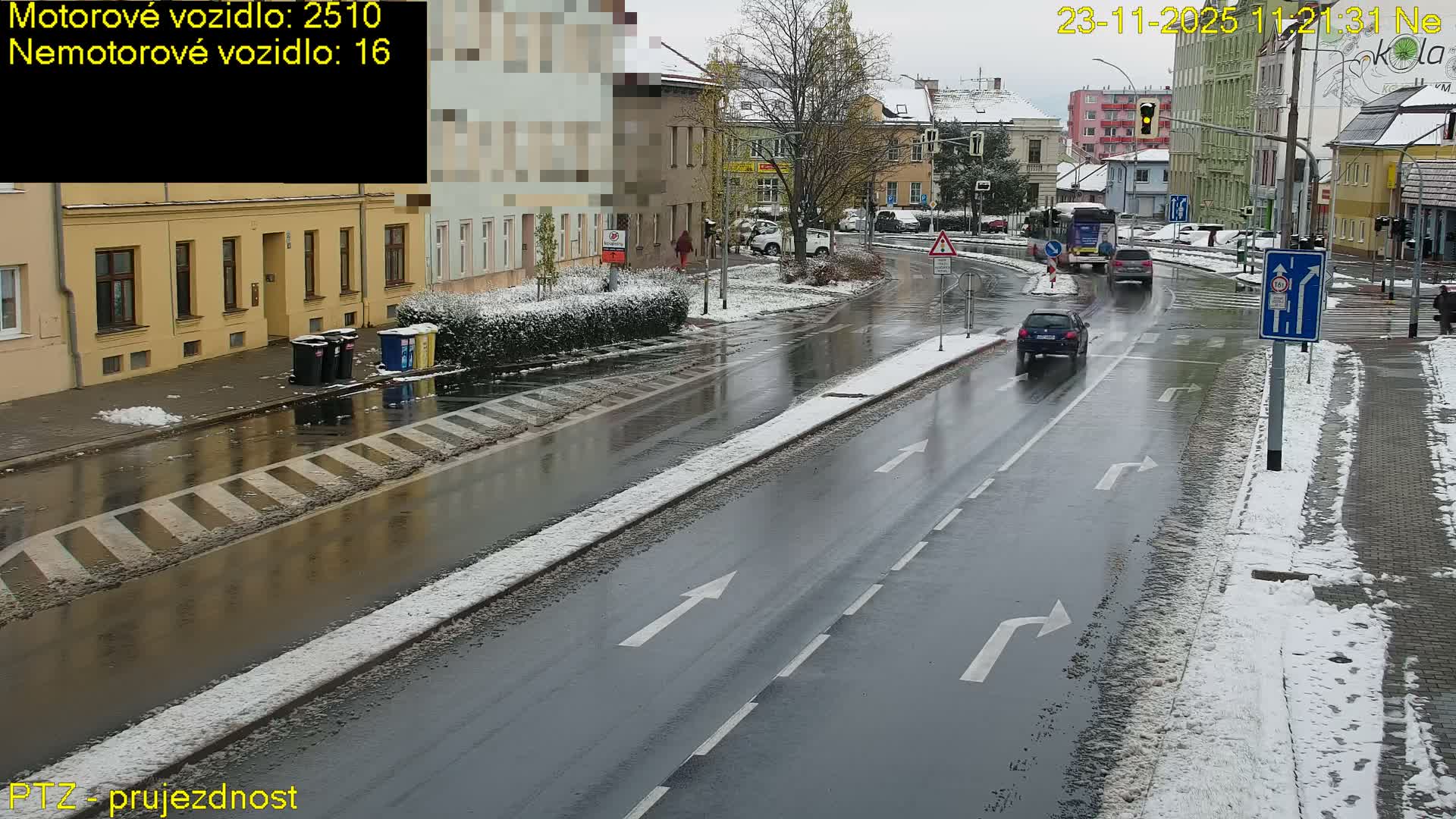 An overcast winter day presents a wet city street edged with snow-covered sidewalks and verges, with cars and a bus in motion alongside buildings and a few pedestrians.