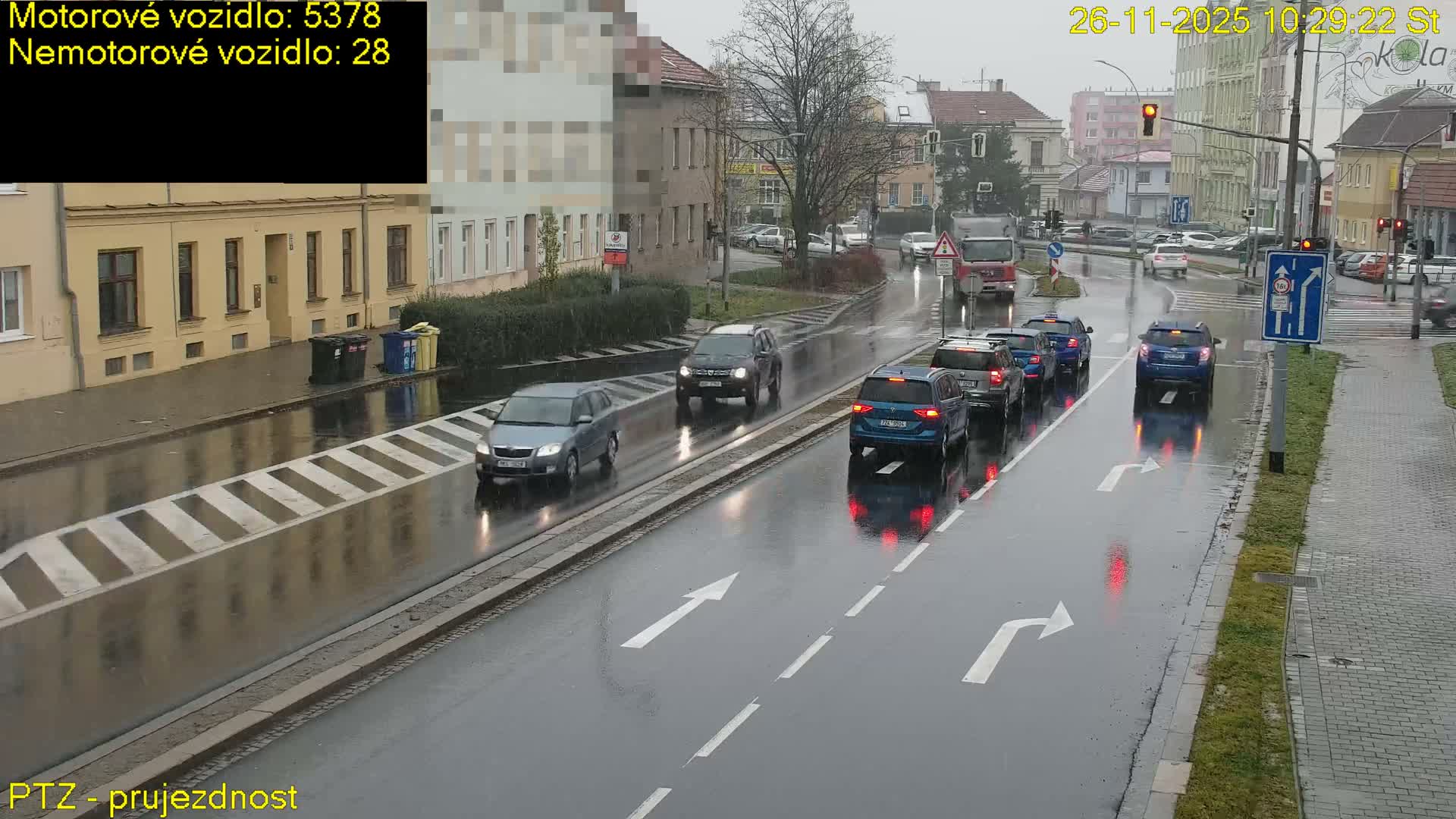 A wet city street is shown from an elevated perspective on an overcast and rainy day, with multiple cars and a truck navigating the road, some queuing at a red traffic light, and buildings lining the background.