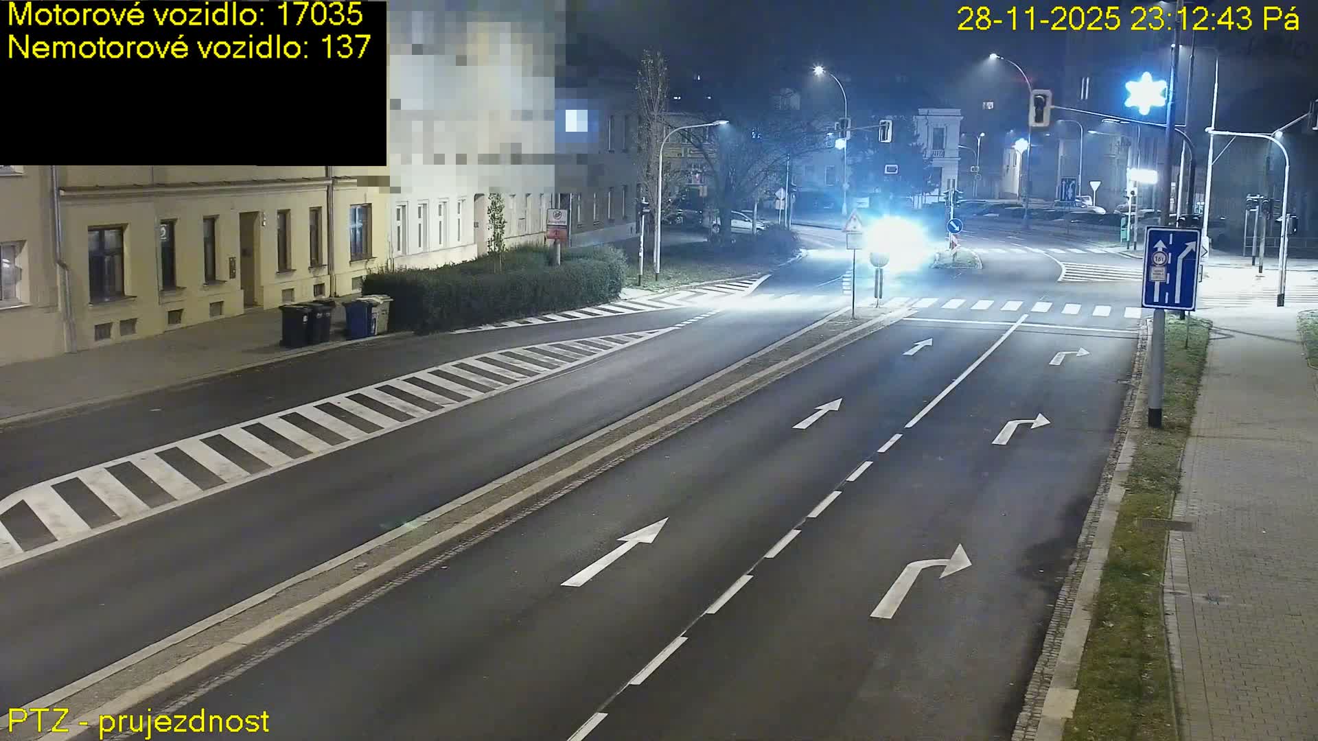 At night, an empty urban intersection with visible road markings and streetlights is illuminated by a car's bright headlights approaching from the right, under clear weather conditions.