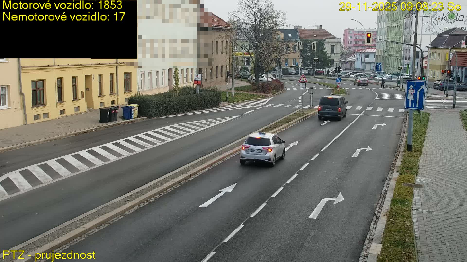 A street scene in a European town depicts an intersection with cars driving, a pedestrian crossing, and buildings under an overcast sky.