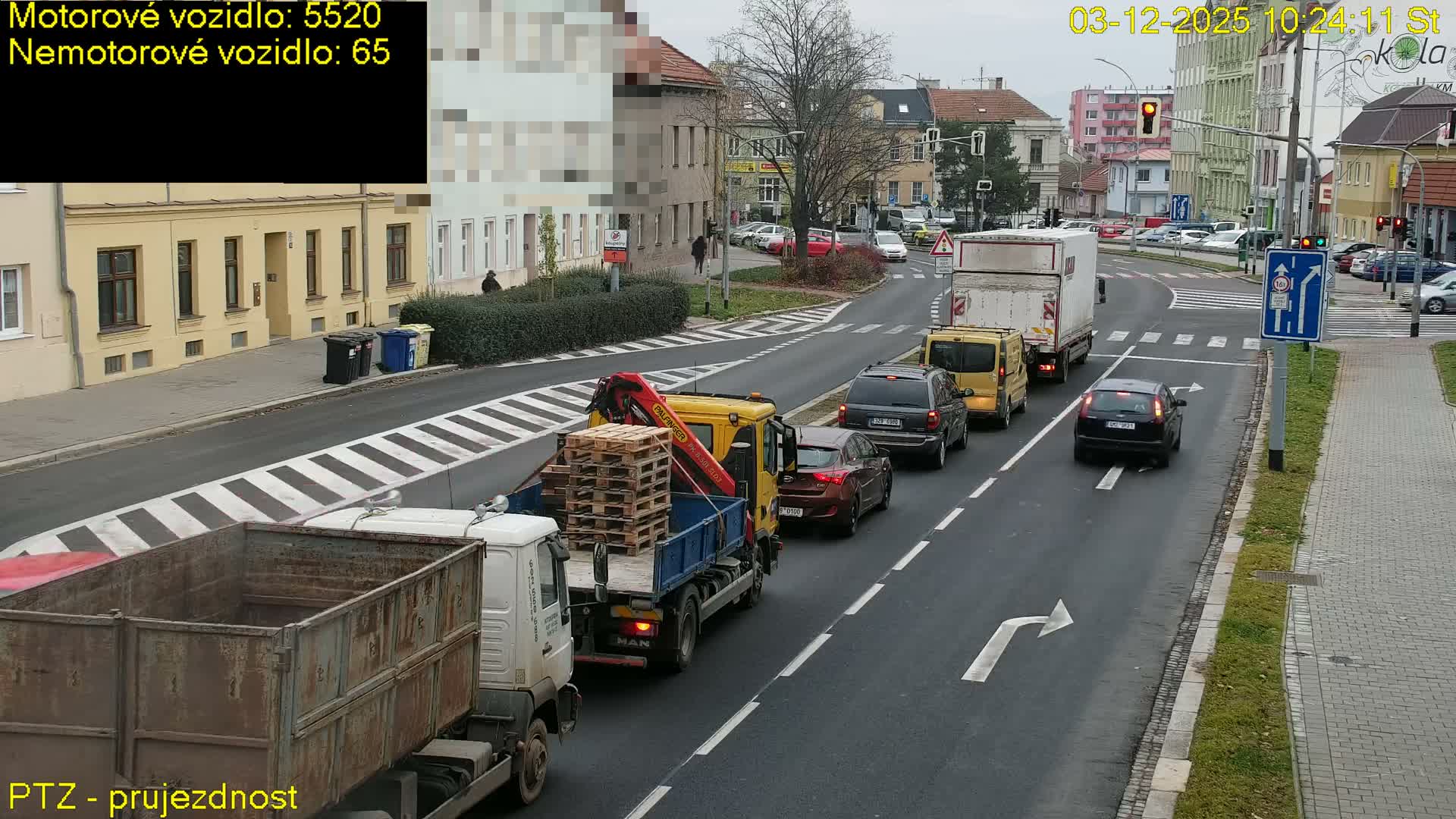 A busy urban street is shown on an overcast day, with various vehicles including cars, vans, and several trucks—one carrying pallets and another with a large skip—navigating an intersection with active traffic lights and pedestrians on the sidewalks.