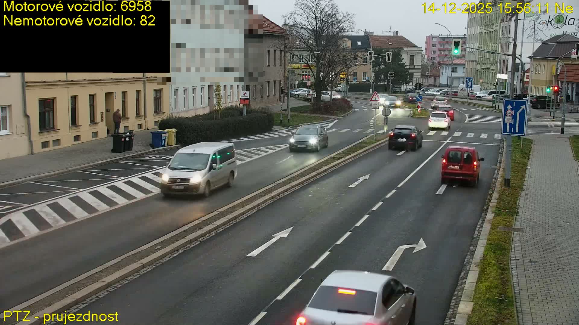 A busy urban street is shown on an overcast day, with various vehicles including cars, vans, and several trucks—one carrying pallets and another with a large skip—navigating an intersection with active traffic lights and pedestrians on the sidewalks.