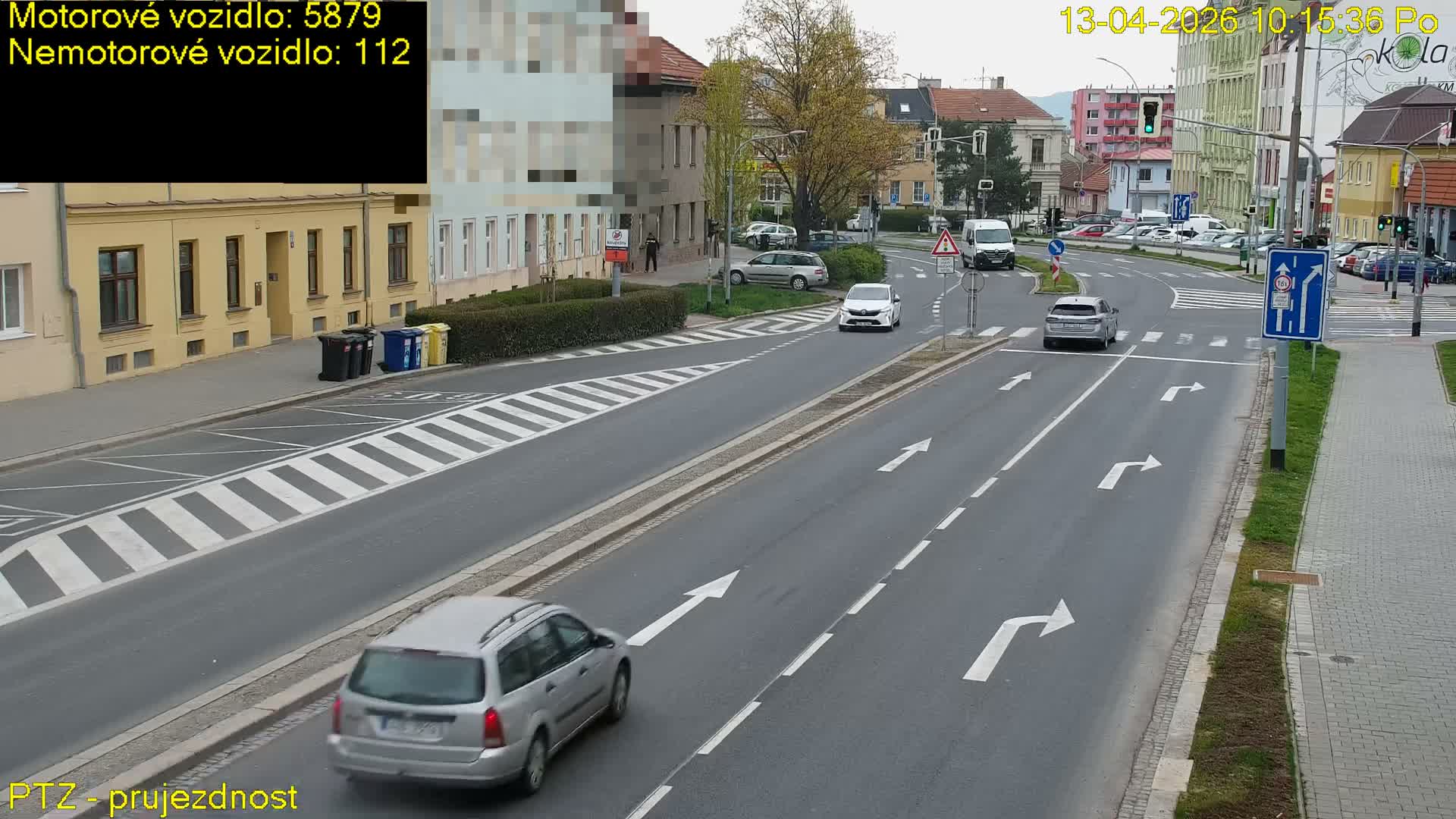 A busy urban street is shown on an overcast day, with various vehicles including cars, vans, and several trucks—one carrying pallets and another with a large skip—navigating an intersection with active traffic lights and pedestrians on the sidewalks.