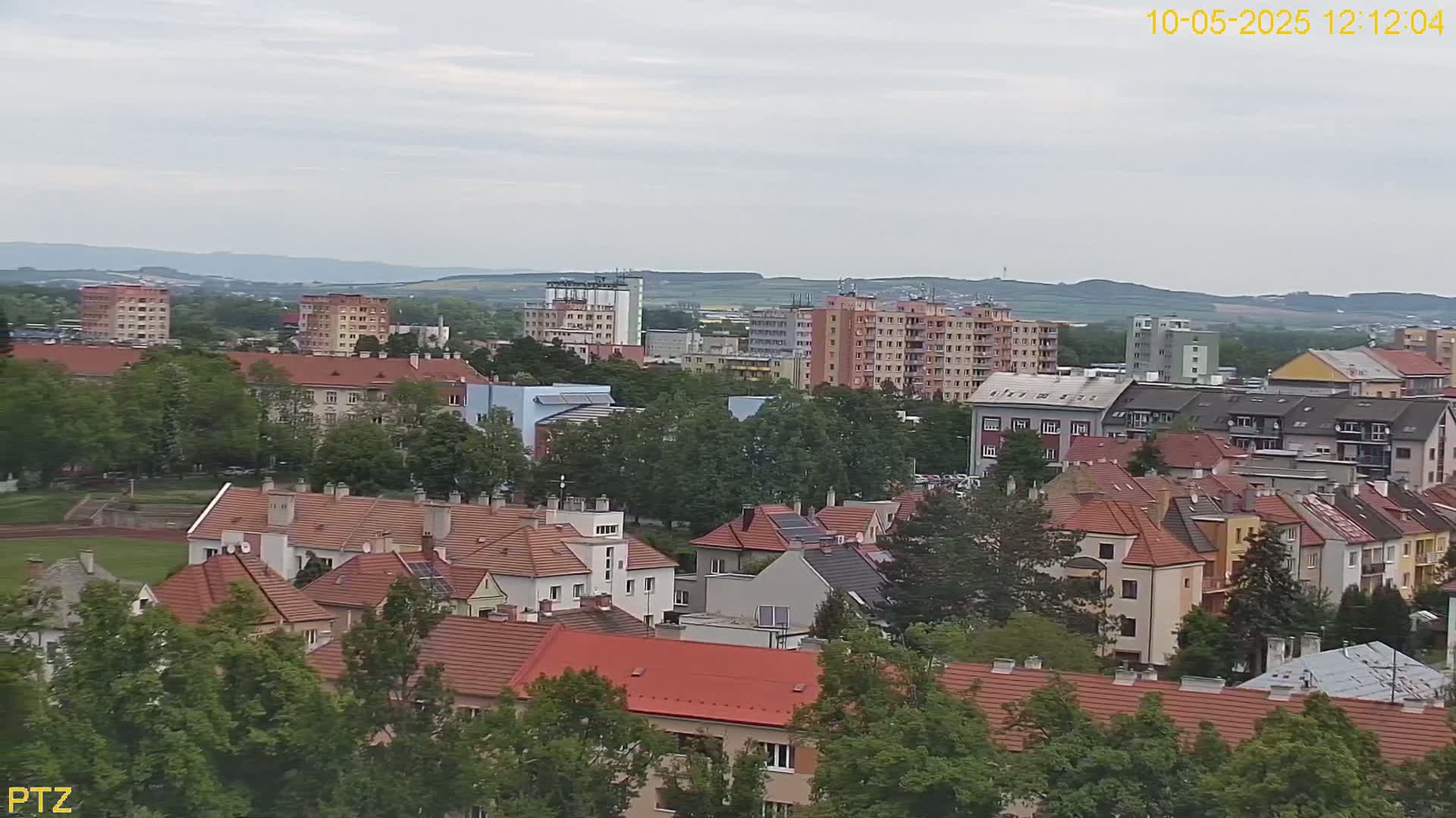A sprawling residential area with red-tiled roofs, interspersed with taller apartment buildings, sits under a cloudy sky, overlooking a grassy field.