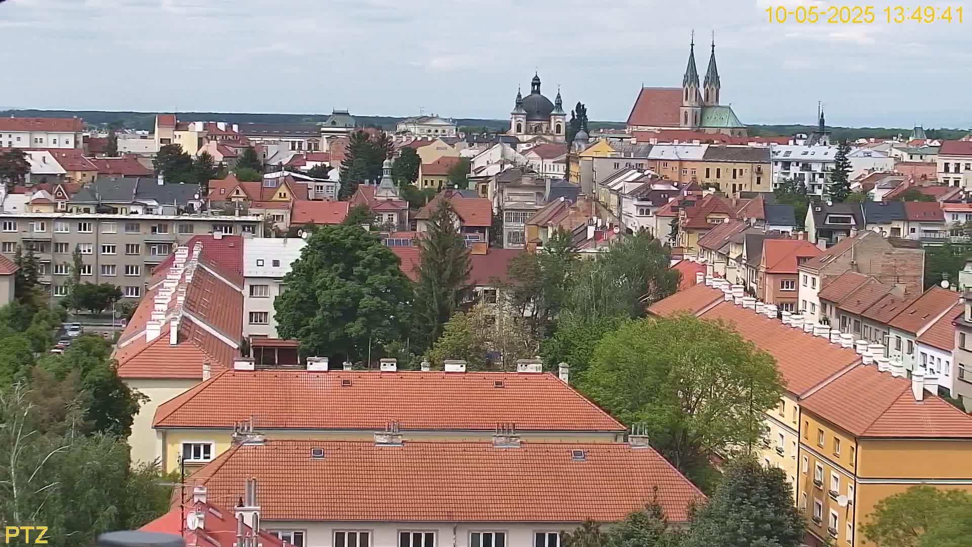 A partly cloudy day reveals a European town with red-tiled roofs, numerous buildings, and a church with twin spires in the distance.