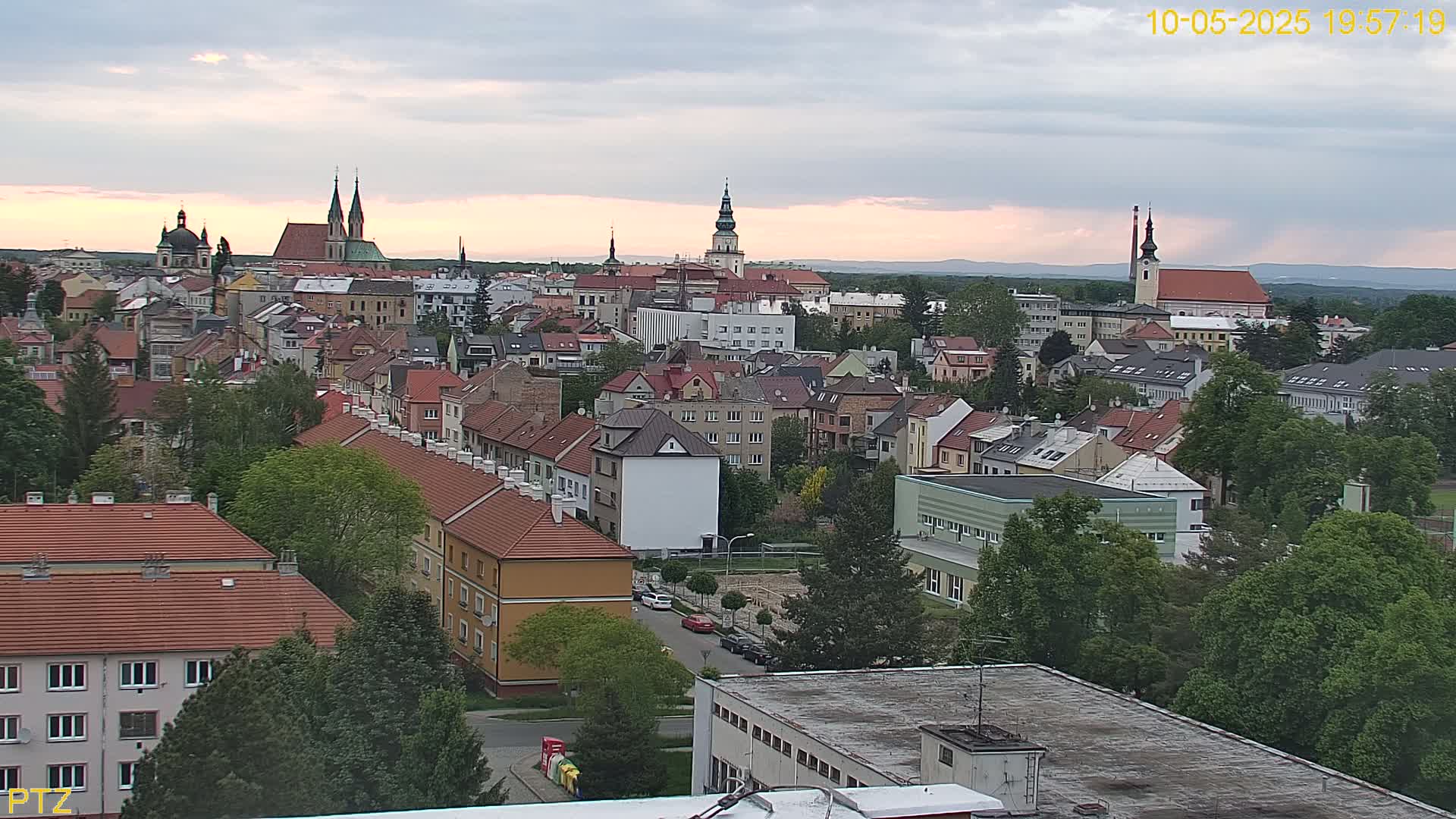 An aerial view of a European town at dusk under a partly cloudy sky shows numerous red-tiled roofs and several church steeples.