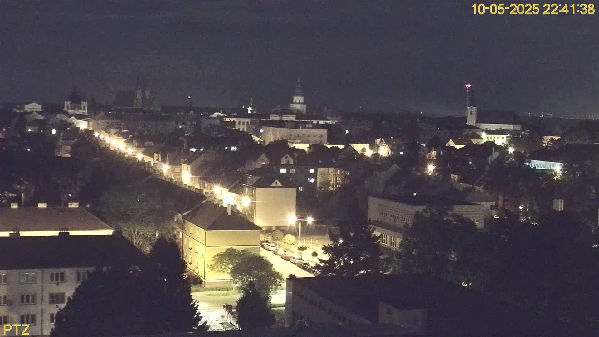A nighttime aerial view of a city, showing a long street lined with lights and buildings extending into the distance under a clear, dark sky.