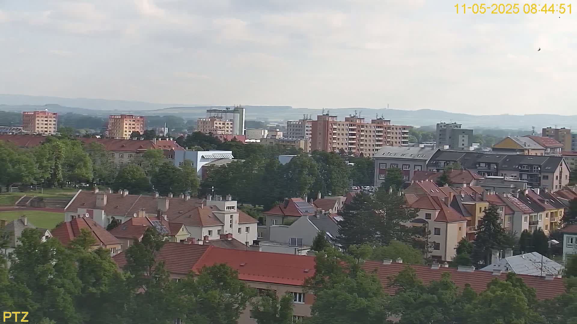 A mostly cloudy day reveals a cityscape of numerous residential buildings with red-tiled roofs, interspersed with taller apartment buildings, and surrounded by lush green trees.