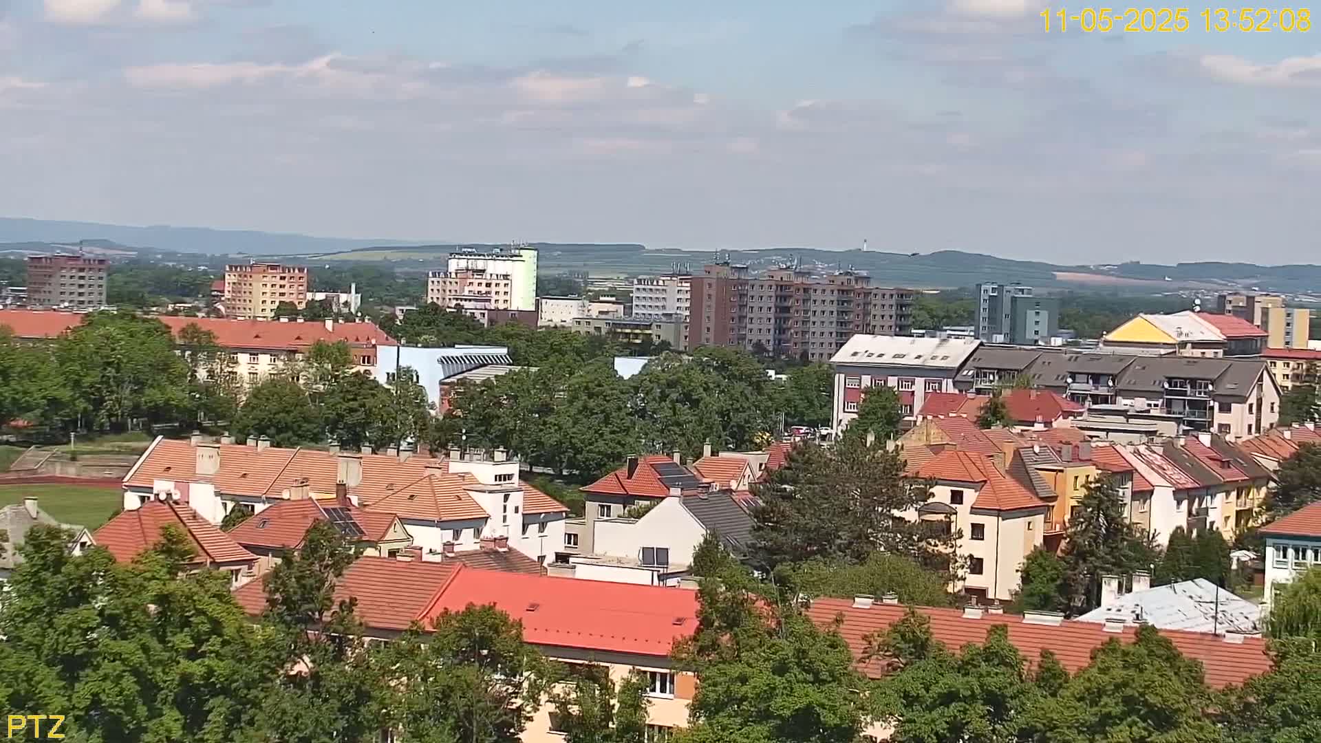 A mostly sunny day reveals a cityscape of many low-rise buildings with red tile roofs, interspersed with taller apartment buildings, and surrounded by lush green trees.