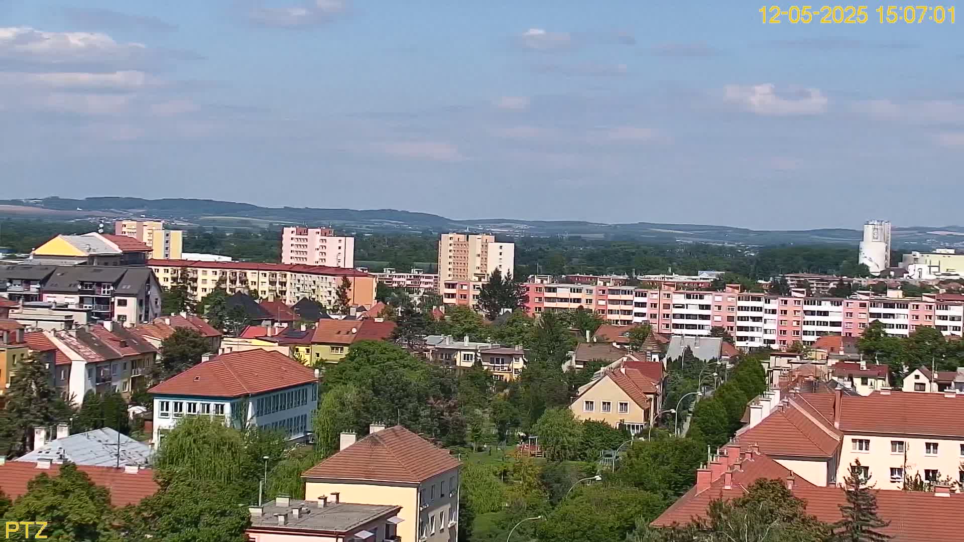 A sprawling town of low-rise buildings and apartment complexes sits nestled amongst lush green trees under a partly cloudy sky.
