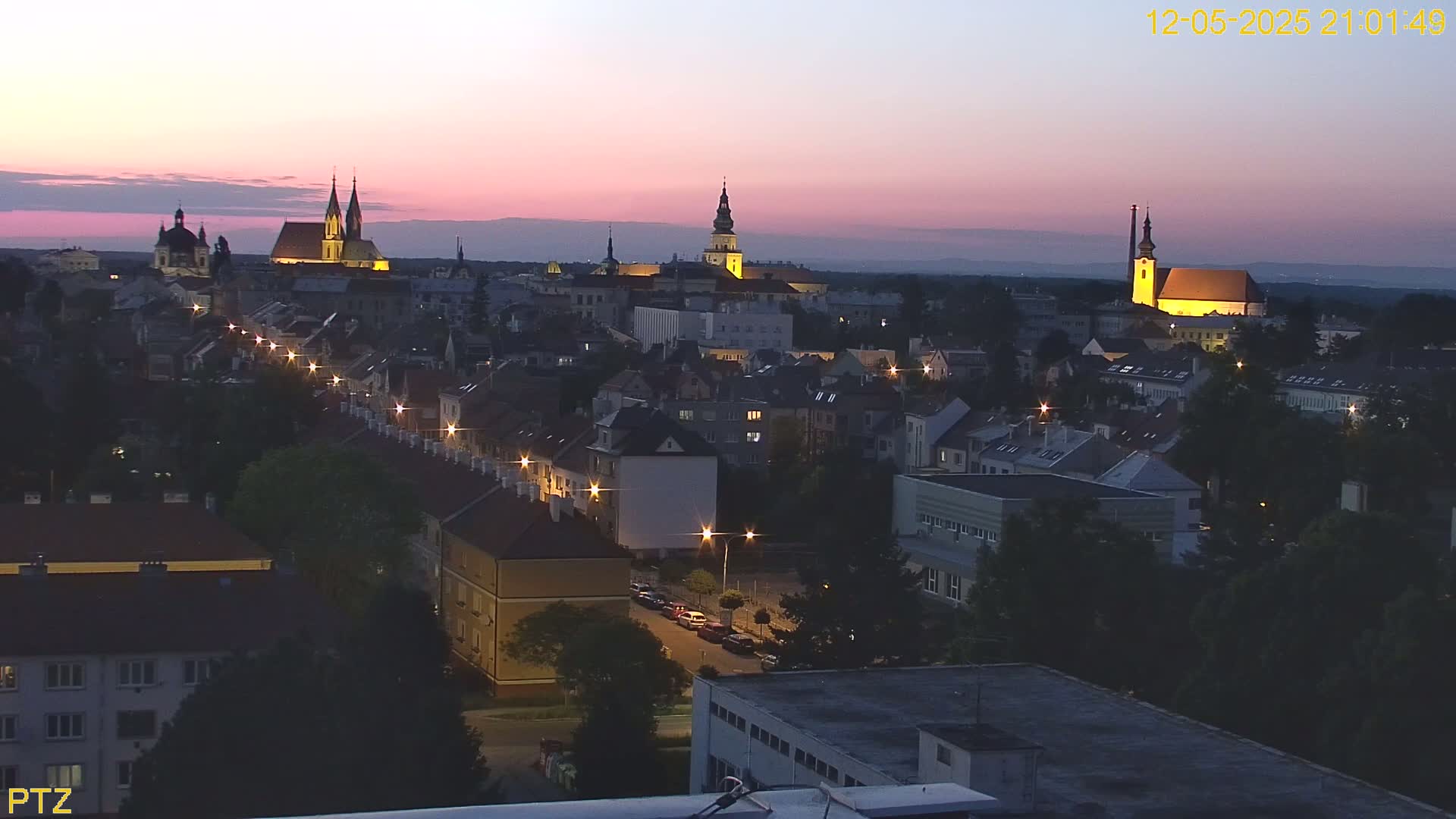 A twilight view of a European city, showing illuminated buildings and streets, with several prominent churches visible in the distance under a pinkish-purple sky.