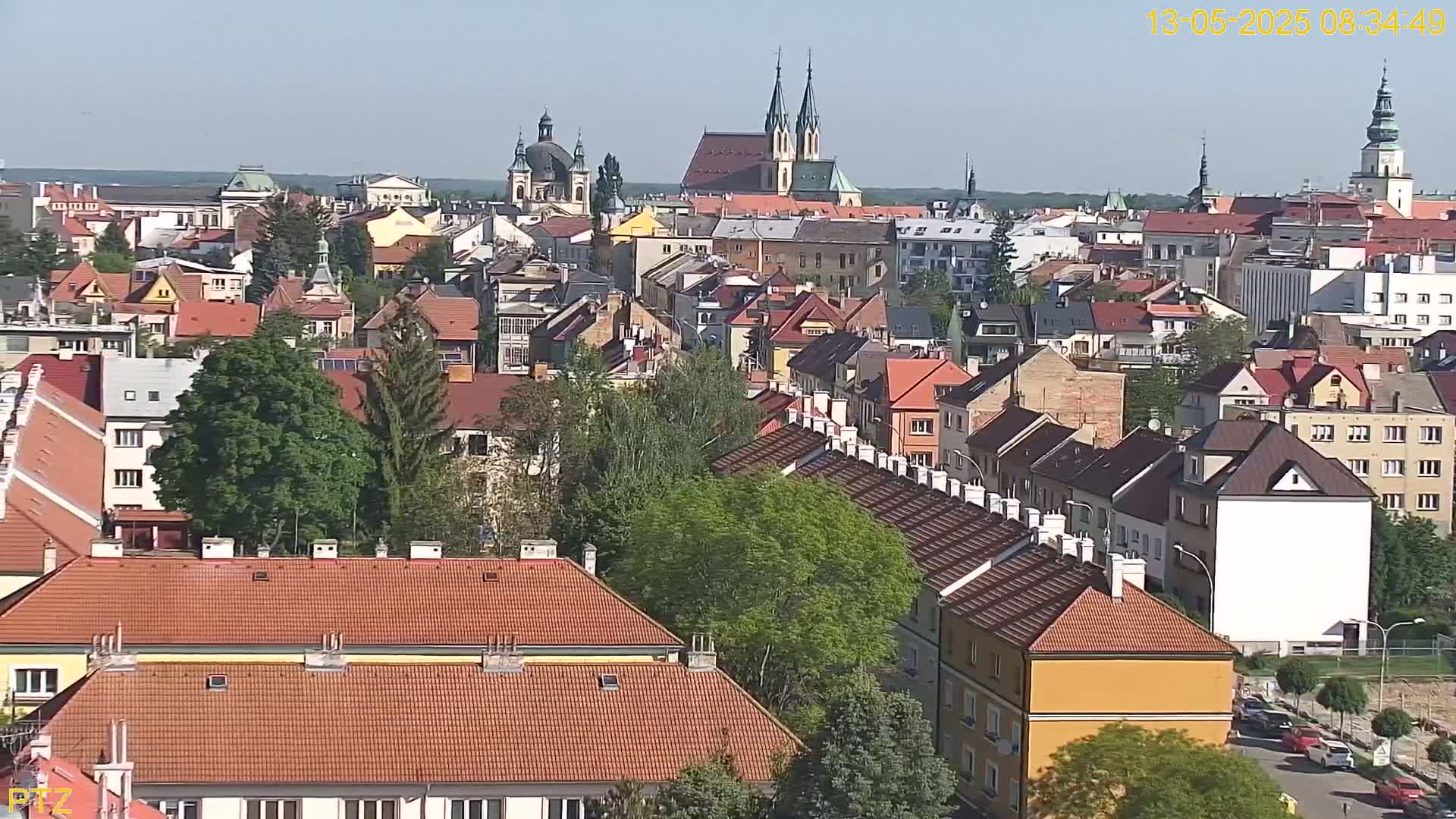 A high-angle, long shot on a sunny day shows a dense collection of red-tiled rooftops of buildings in a European city, with several church steeples visible in the distance.