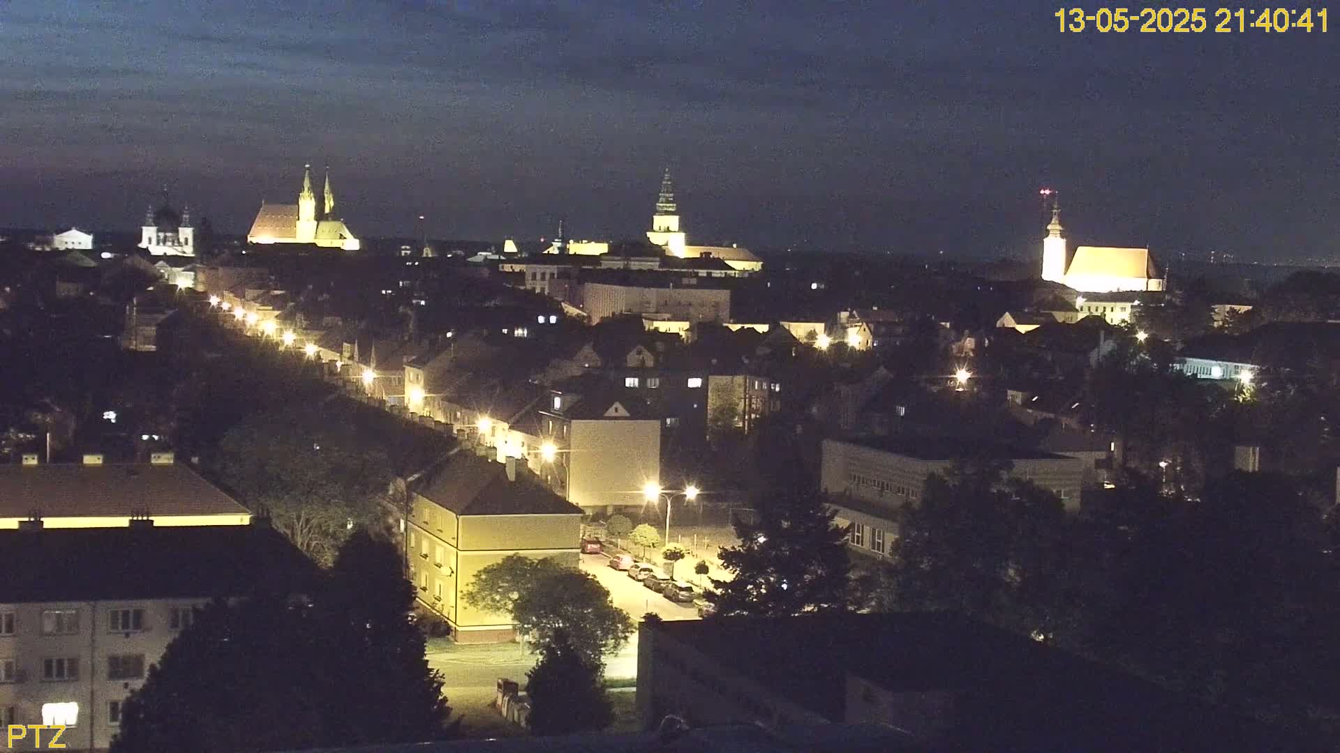 A nighttime, high-angle view of a European town with illuminated buildings and streets, featuring several prominent churches or cathedrals.