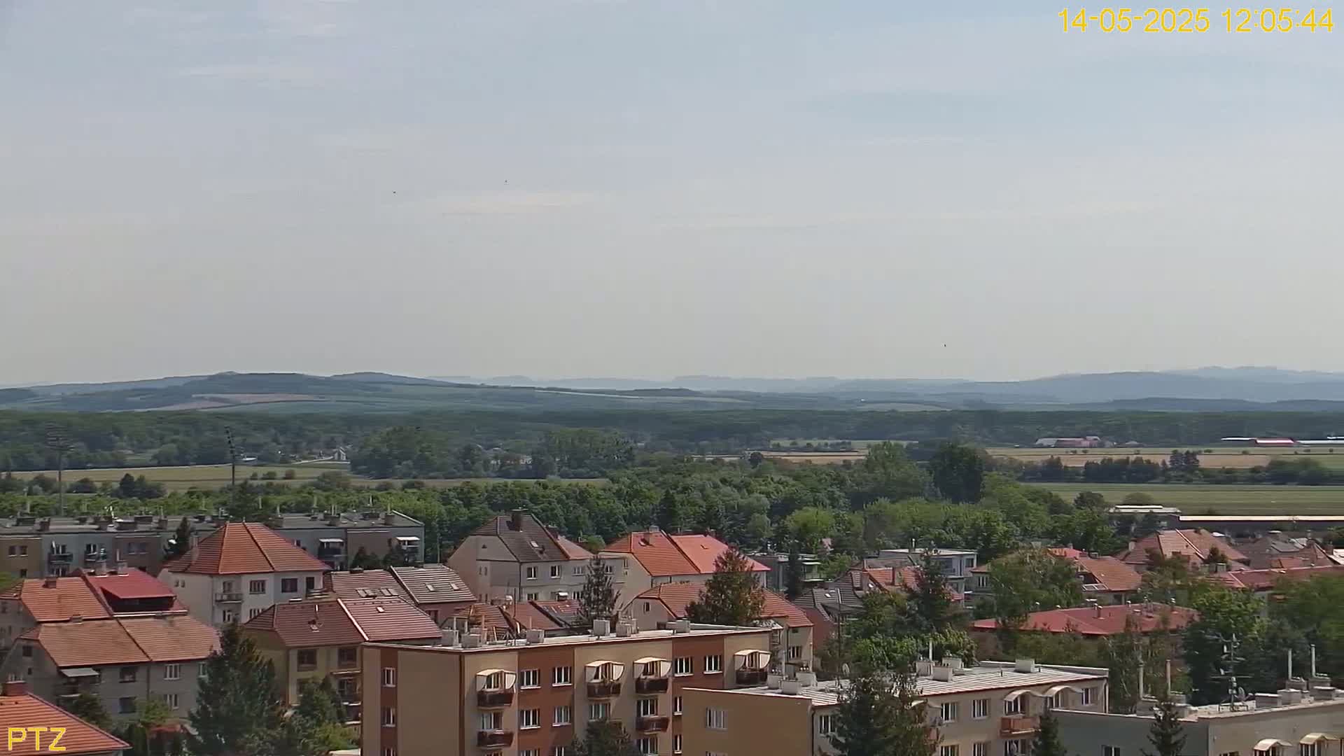 A hazy, sunny day overlooks a town of red-roofed buildings nestled beside a verdant landscape of trees and rolling hills.