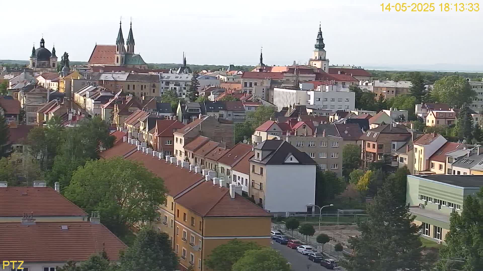 A mostly sunny view of a European town with many red-tiled roofs, several churches, and green trees, seen from a high vantage point.