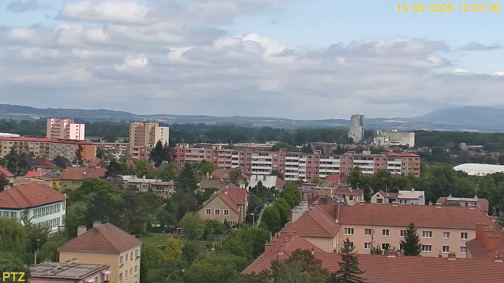 A partly cloudy day reveals a sprawling cityscape of multi-story apartment buildings and smaller residential houses, with a distant view of hills and industrial structures.