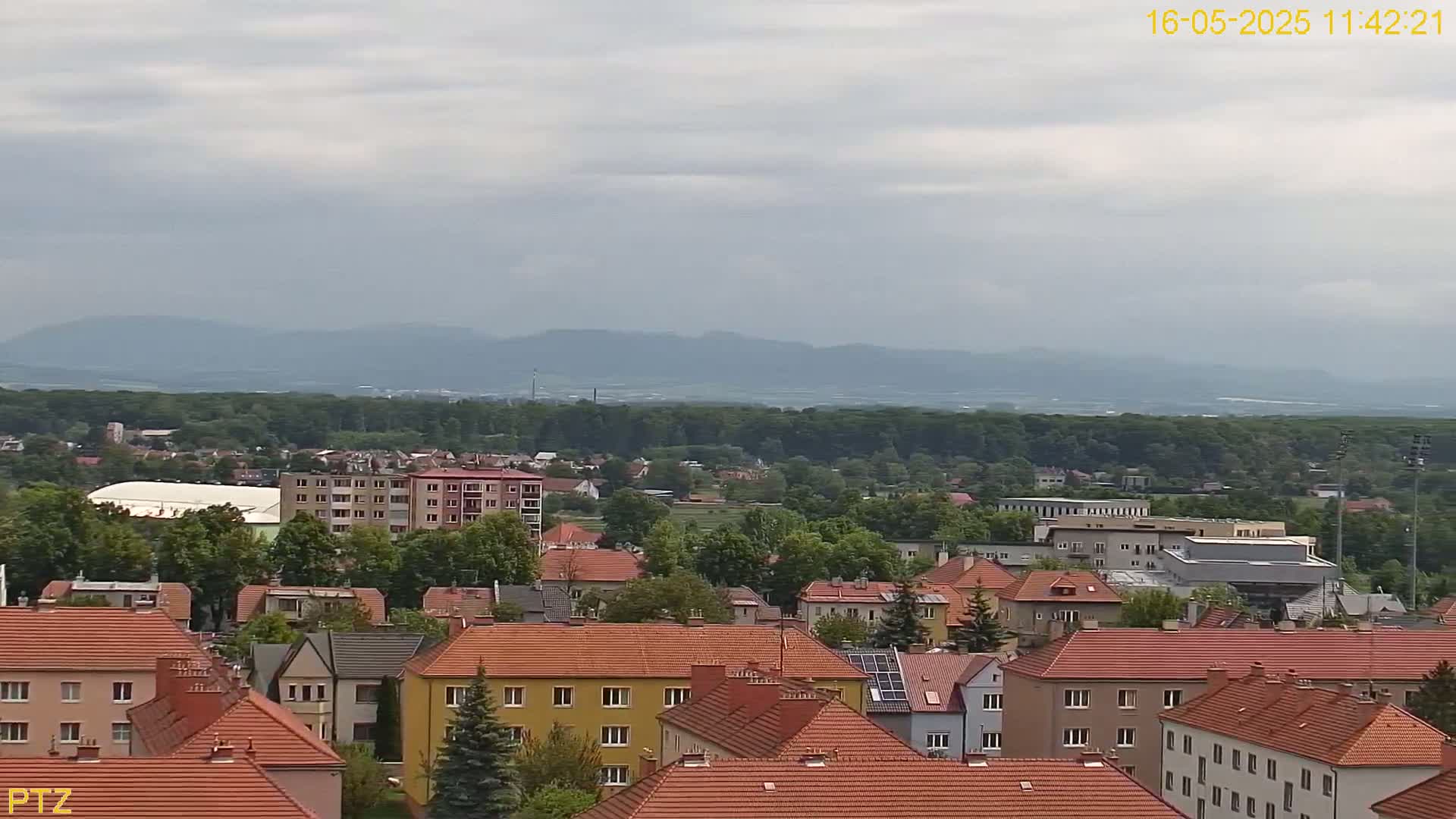 A view of a town with red-tiled roofs, surrounded by green trees and distant mountains under a cloudy sky.