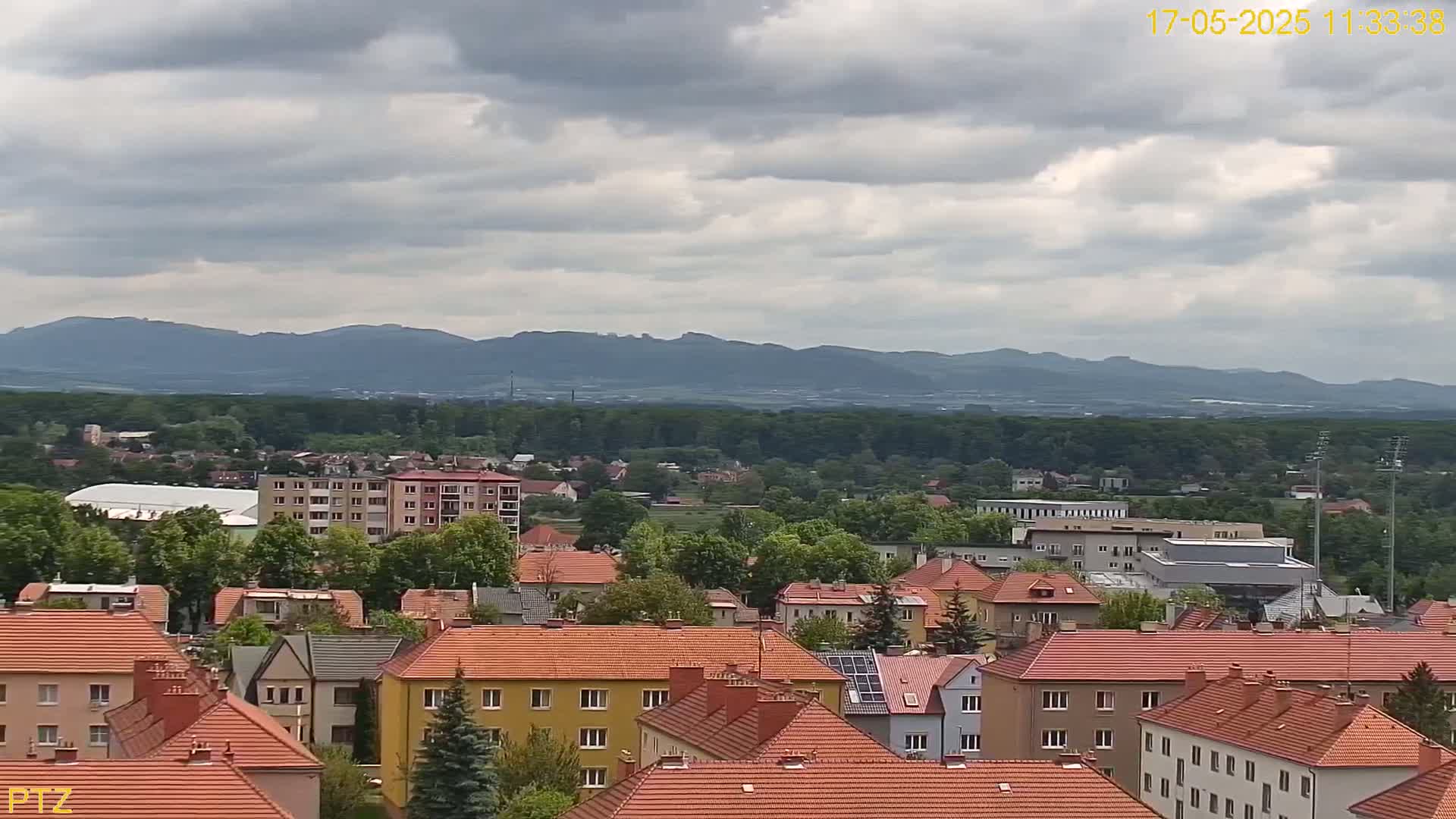 A town of red-roofed buildings sits in a valley under a cloudy sky, with mountains visible in the distance.