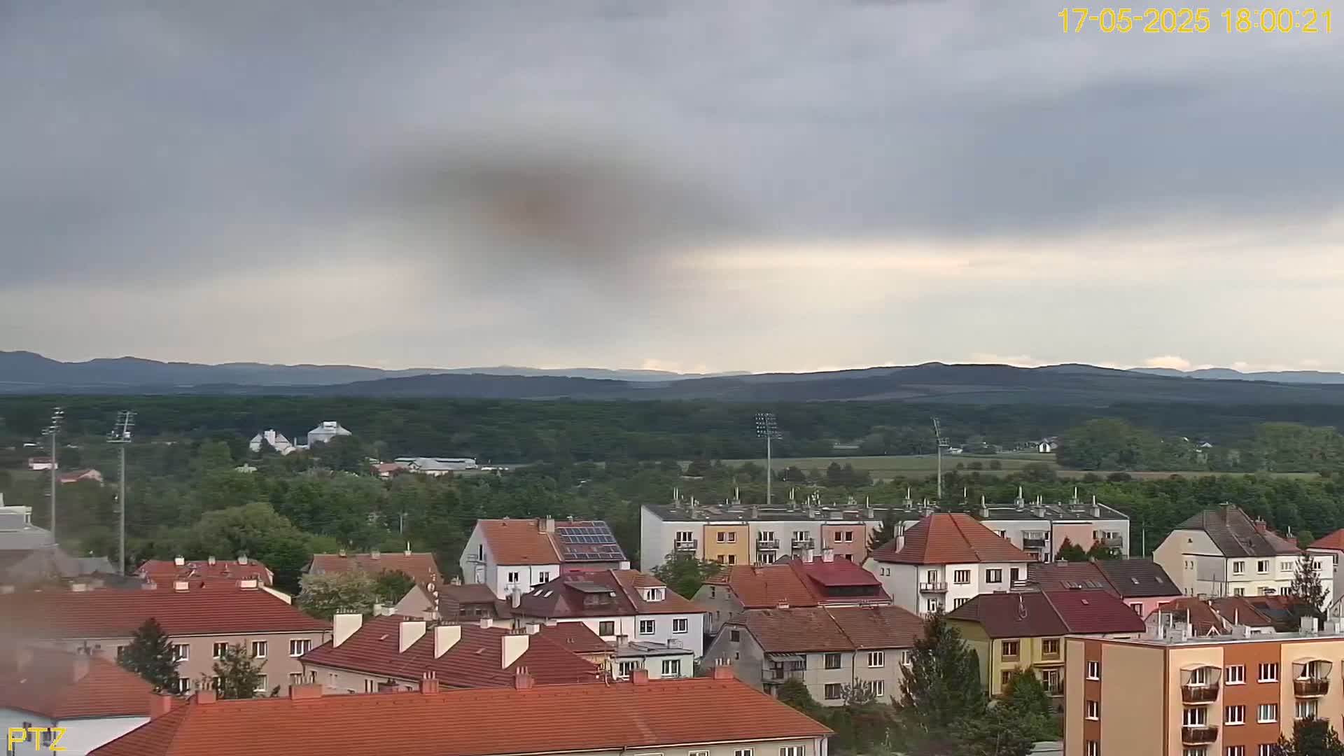 A view of a town with red-tiled roofs, under a mostly cloudy sky, shows a range of low hills in the distance.