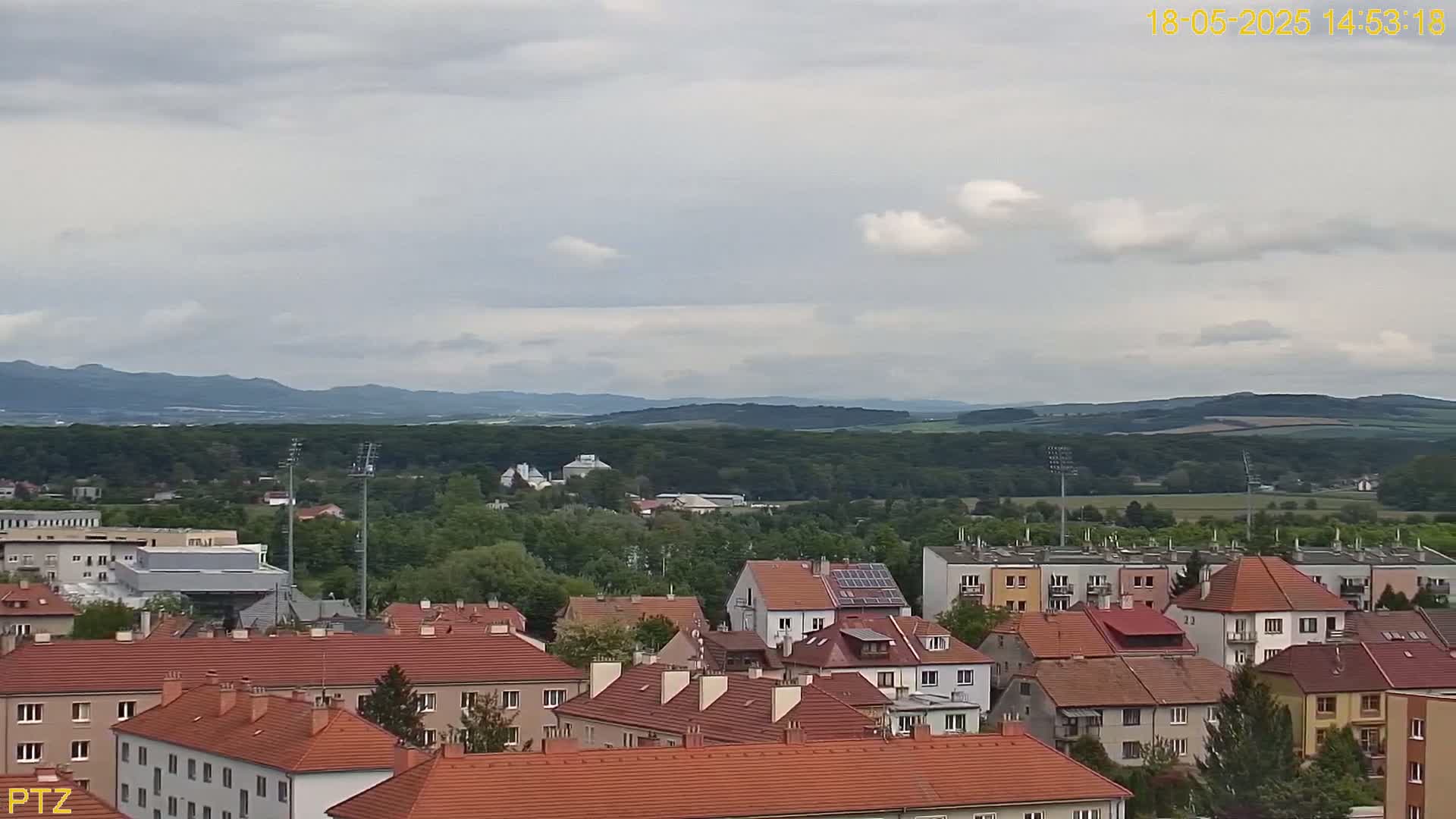 An overcast day reveals a sprawling town of red-roofed buildings nestled against a backdrop of lush green hills and distant mountains.