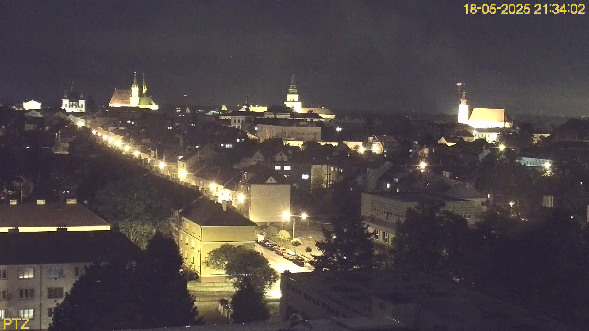 A nighttime aerial view of a town shows illuminated buildings and streets, with several prominent church spires visible in the distance.