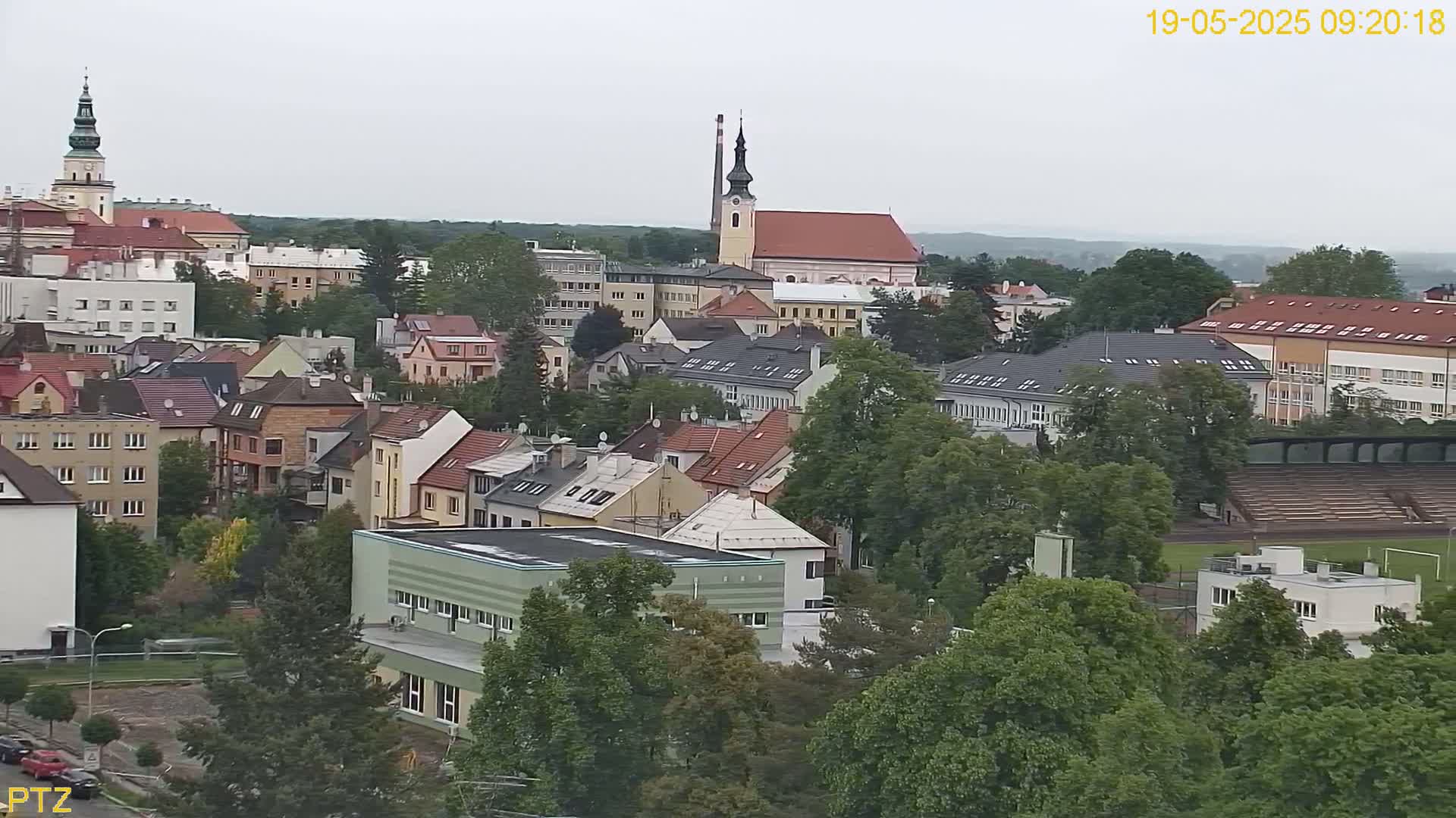 An aerial view of a town on an overcast day, showing a mix of residential buildings, a church steeple, and a stadium nestled among lush green trees.