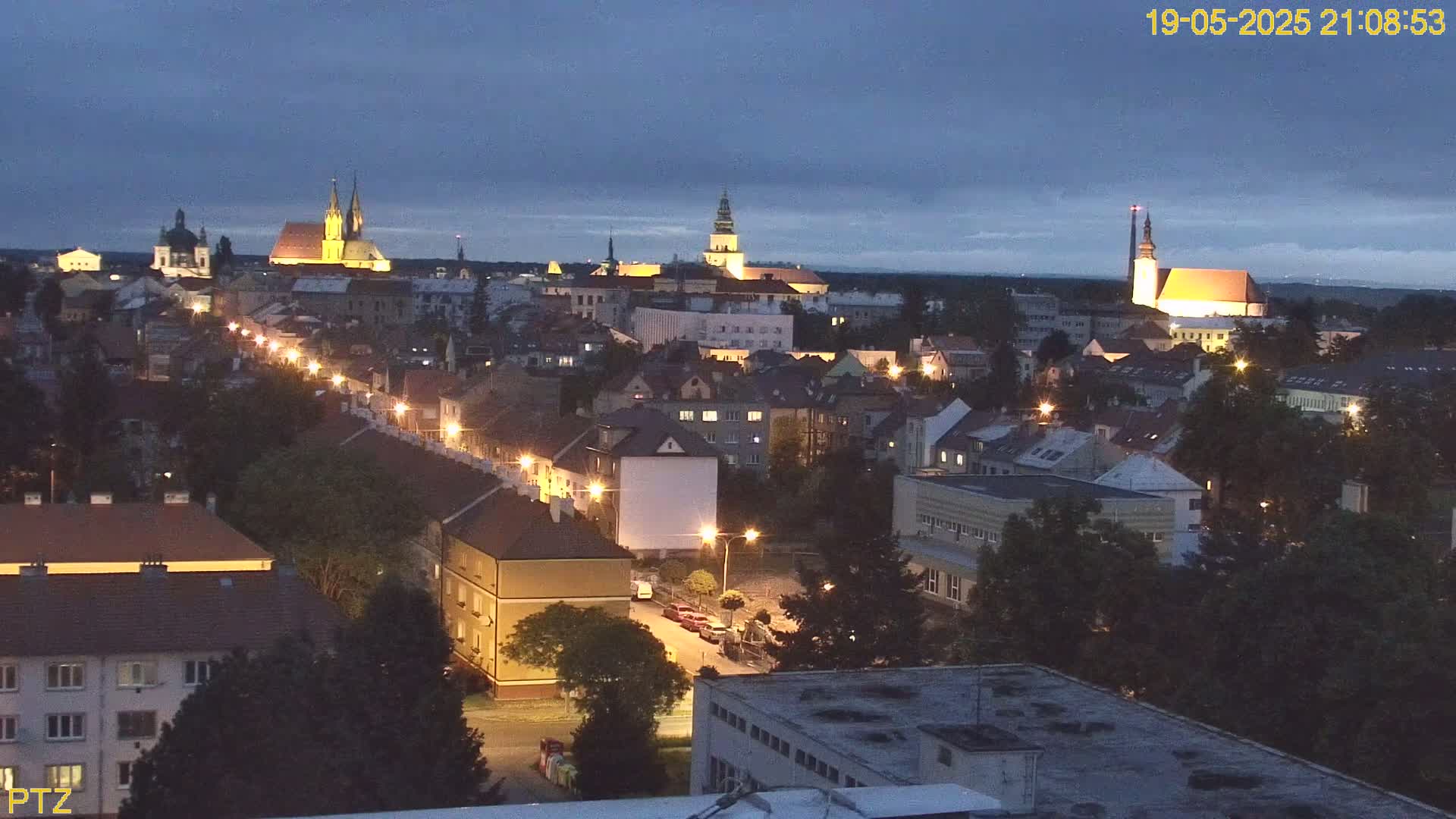 A nighttime view of a European city with illuminated buildings and streets, under a partly cloudy twilight sky.