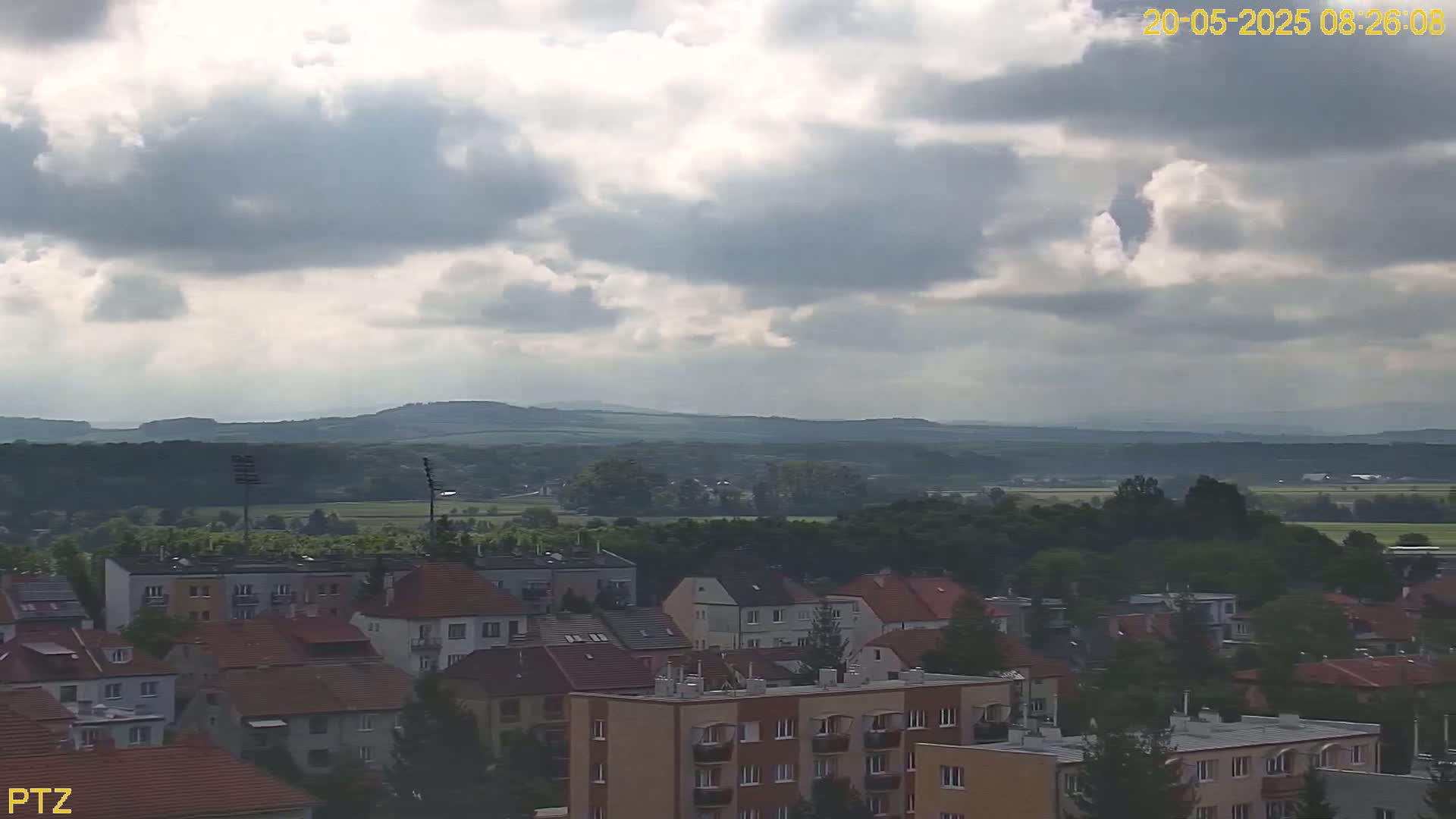 A partly cloudy sky overlooks a town of low-rise buildings nestled against a line of distant, rolling hills.