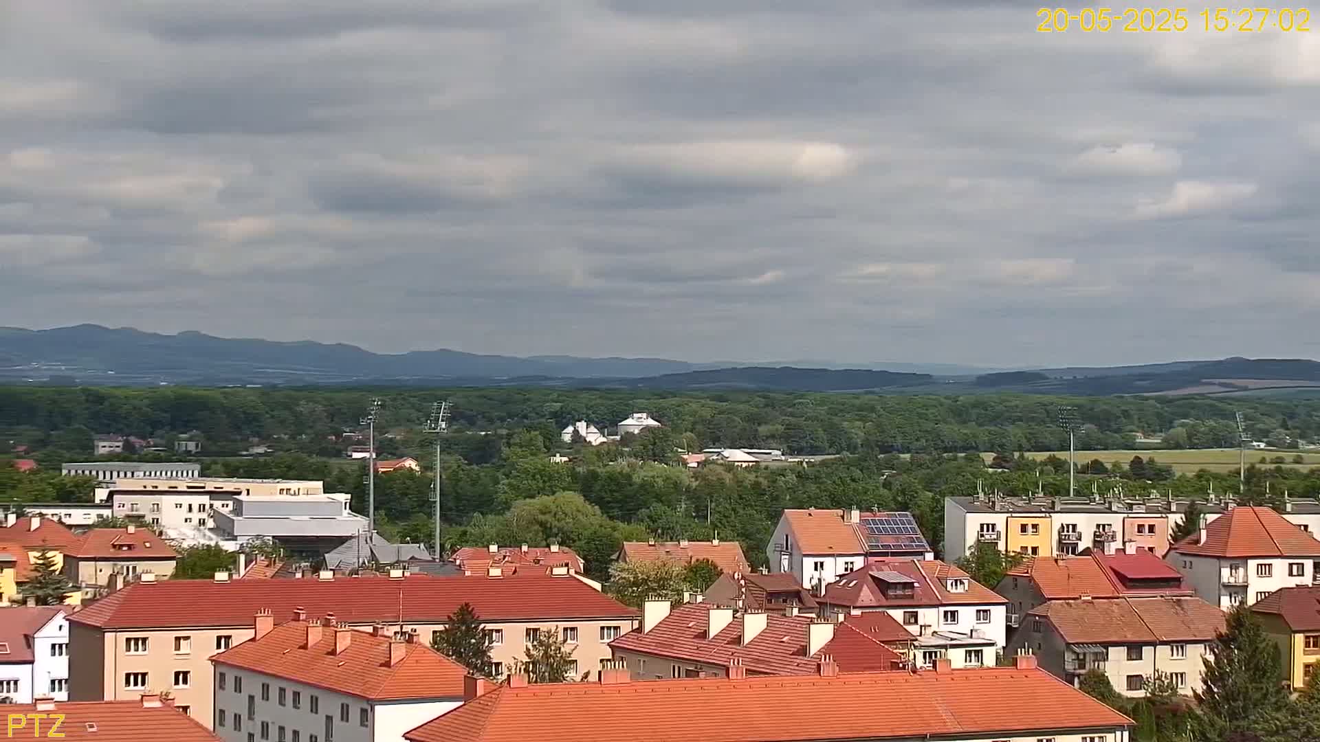An aerial view shows a town of red-roofed houses under a mostly cloudy sky, with a line of distant hills in the background.
