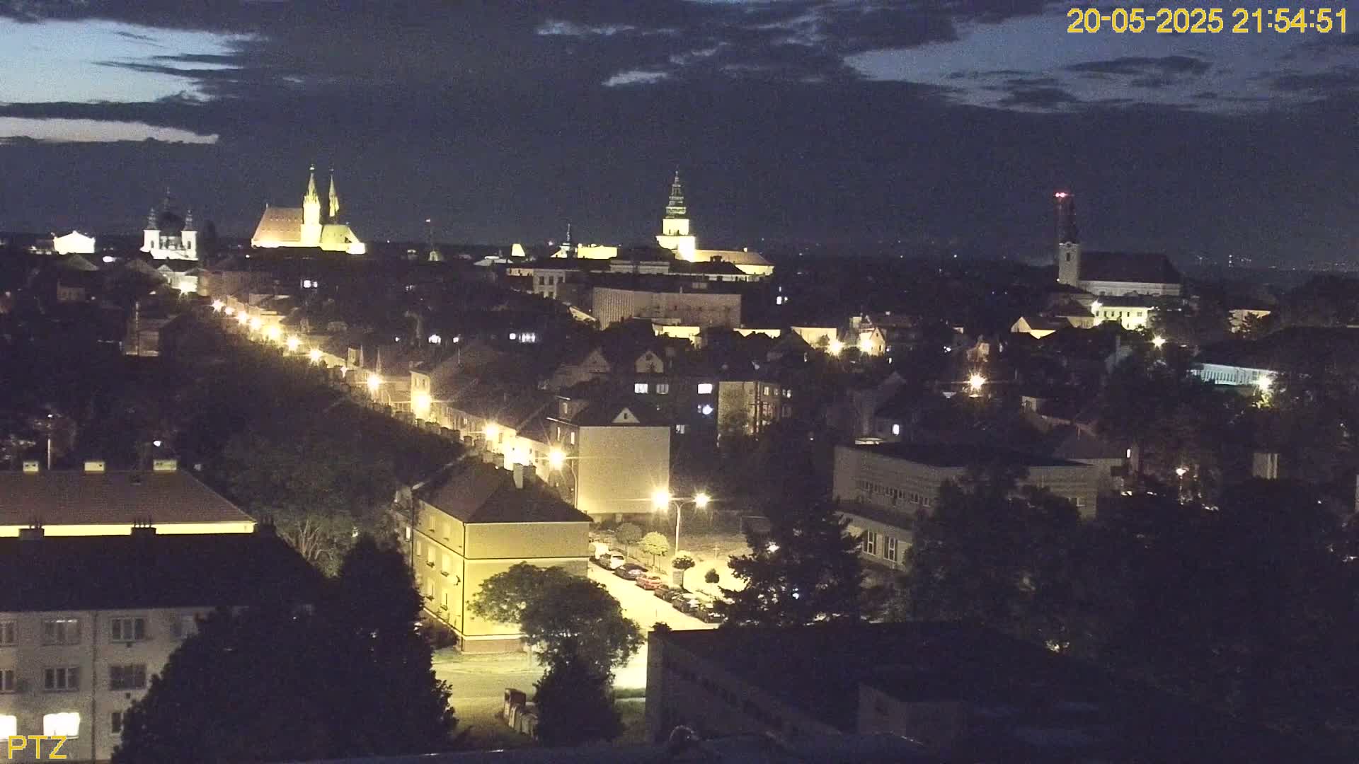 A nighttime, high-angle view of a town illuminated by streetlights, with several churches visible in the distance under a partly cloudy sky.