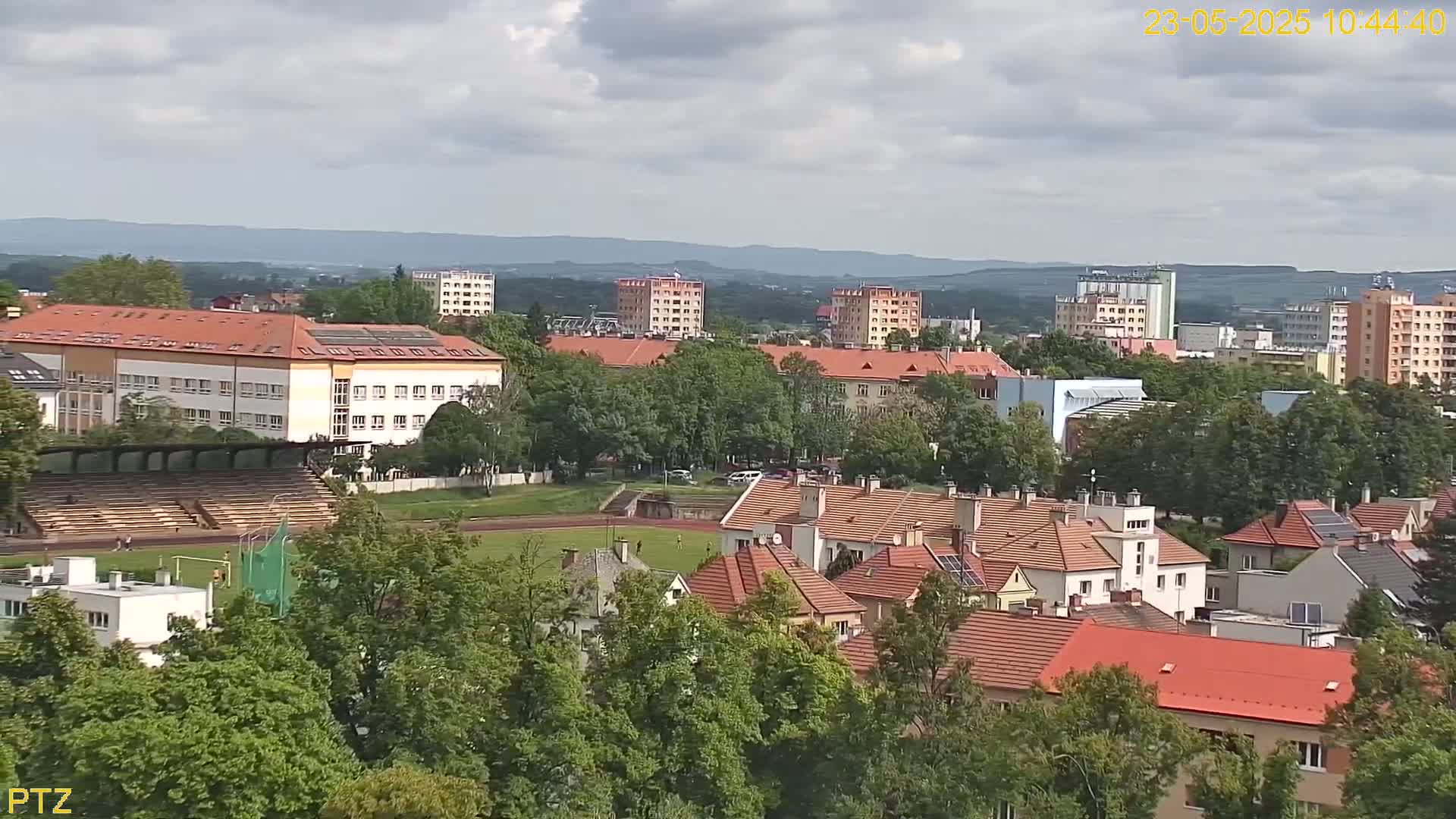 An elevated view shows a town under a partly cloudy sky, with a large school building, a track and field stadium, and numerous residential buildings with red-tiled roofs surrounded by lush green trees.