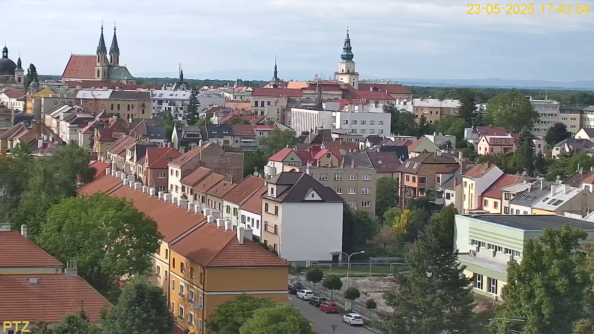 A high-angle view of a European town on a partly cloudy day, showing many red-tiled roofs and several prominent church steeples in the distance.