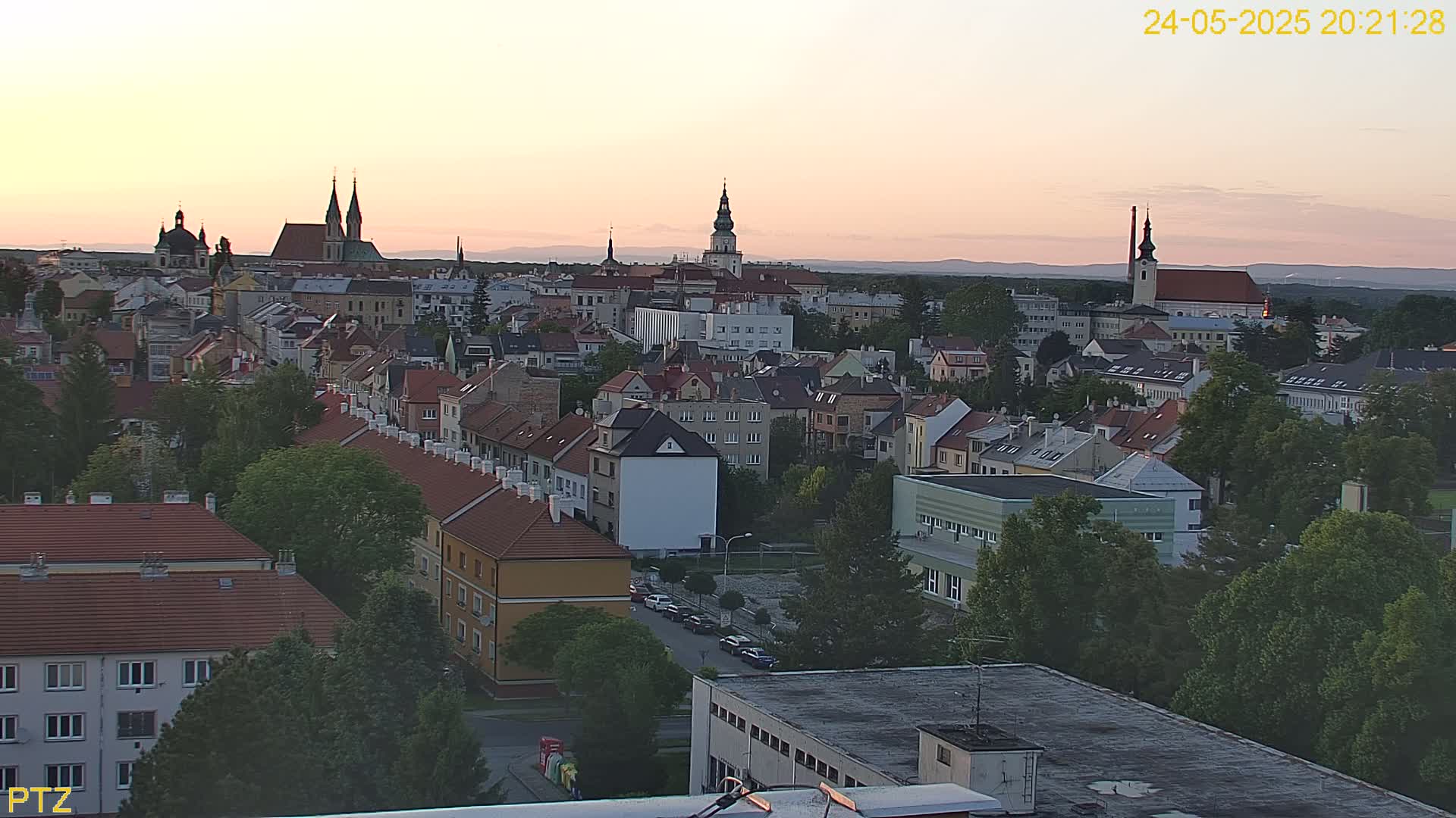 An elevated view of a European city at sunset, showing a dense collection of buildings with red-tiled roofs, interspersed with green trees.