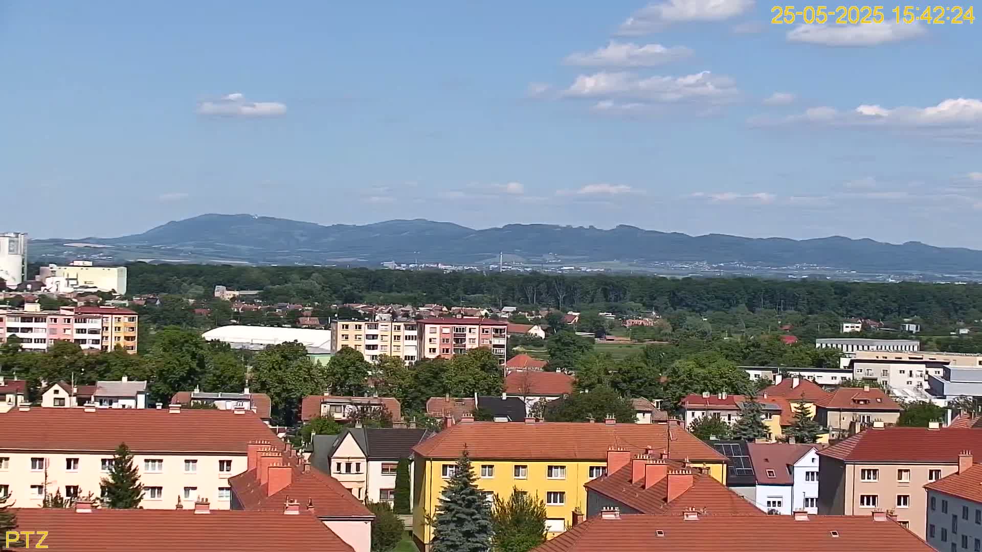 A mostly sunny day reveals a residential area with red-tiled roofs, nestled among green trees, with a distant mountain range visible on the horizon.