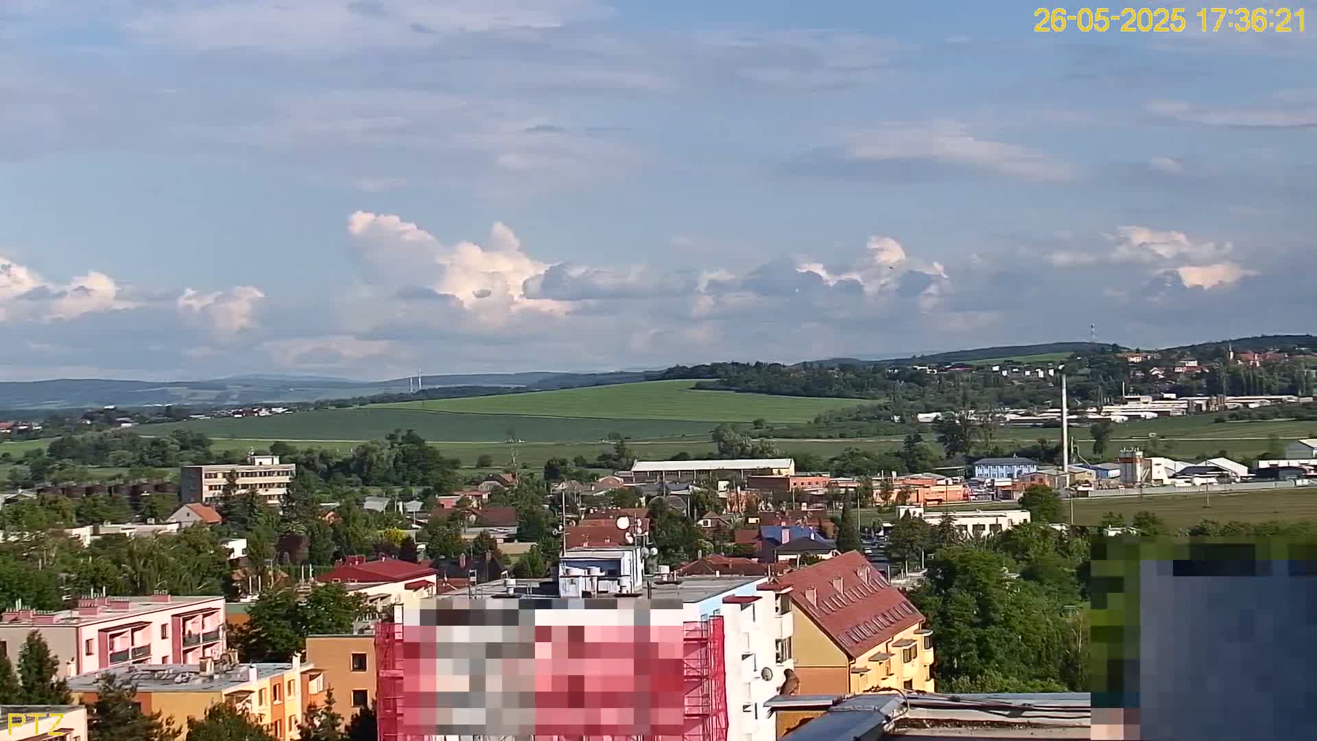 A partly cloudy daytime view of a town nestled in a valley, with rolling green hills and distant mountains in the background.