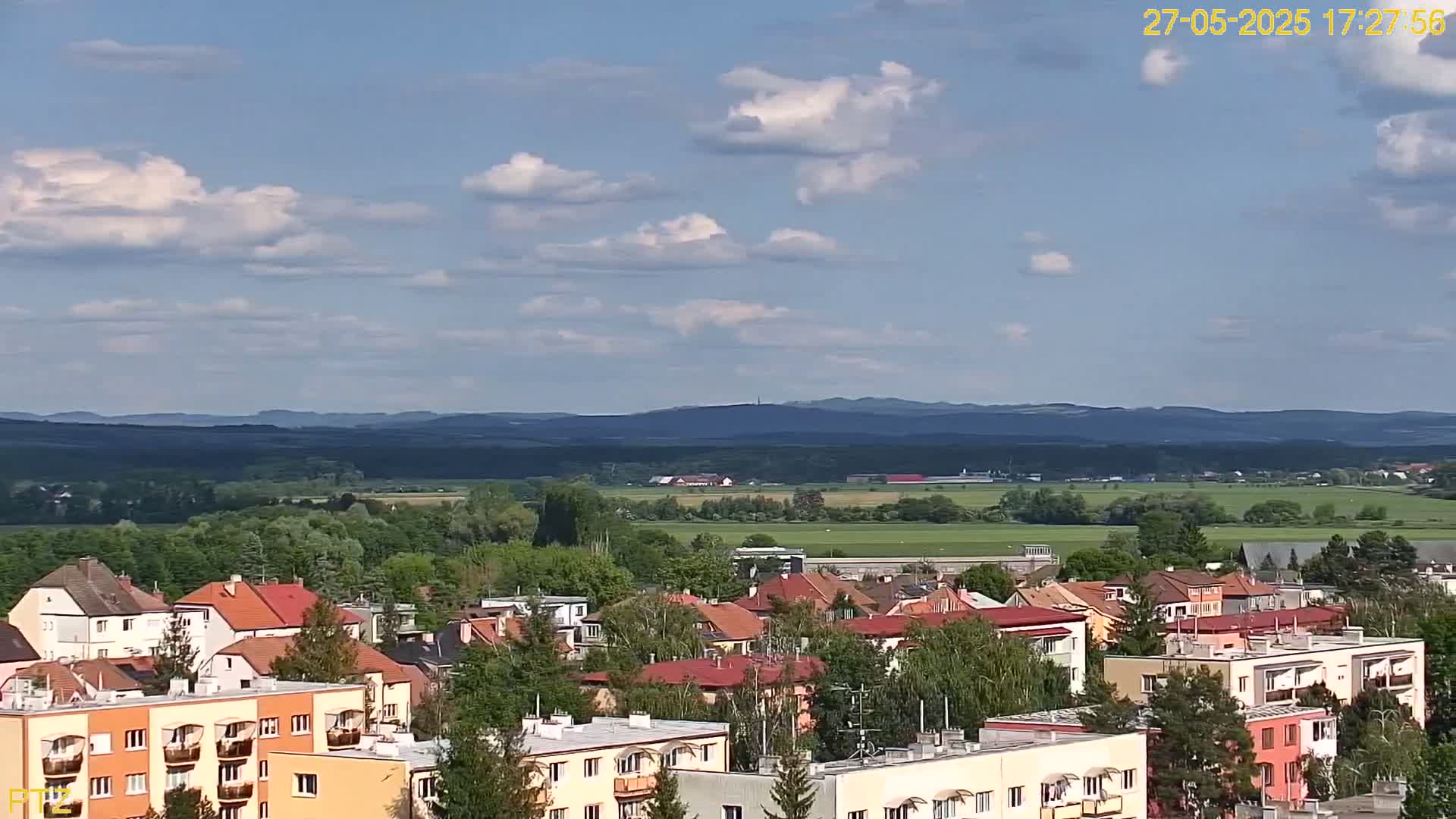 A partly cloudy day reveals a view of a town nestled amongst green trees and fields, with distant mountains under a mostly blue sky.