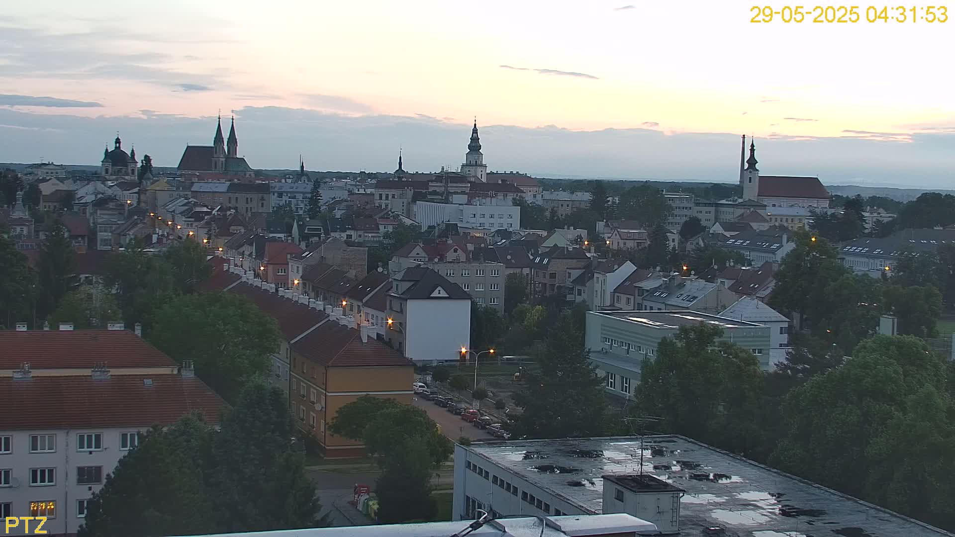 An elevated view of a European city at twilight, showing numerous buildings, trees, and several church steeples, under a mostly clear sky.