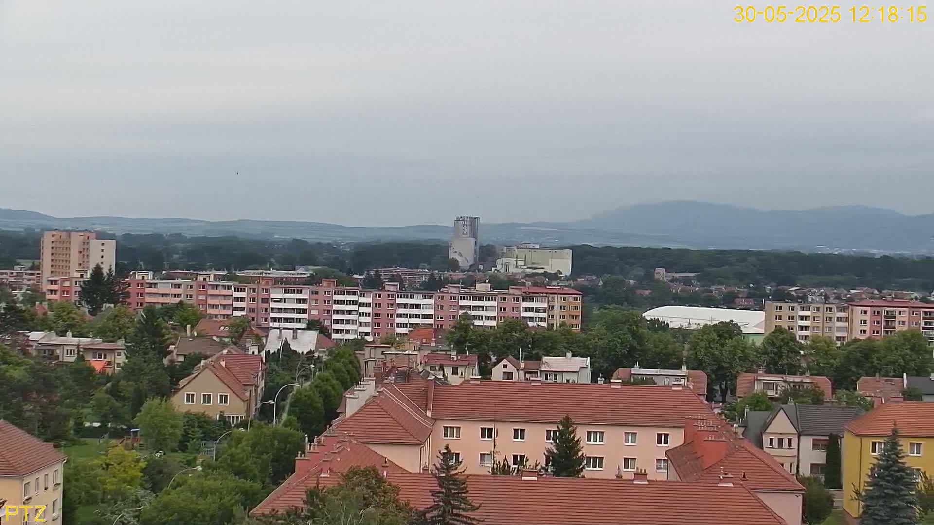 A sprawling residential area with red-tiled roofs, apartment buildings, and trees, sits under an overcast sky, with distant mountains visible in the background.