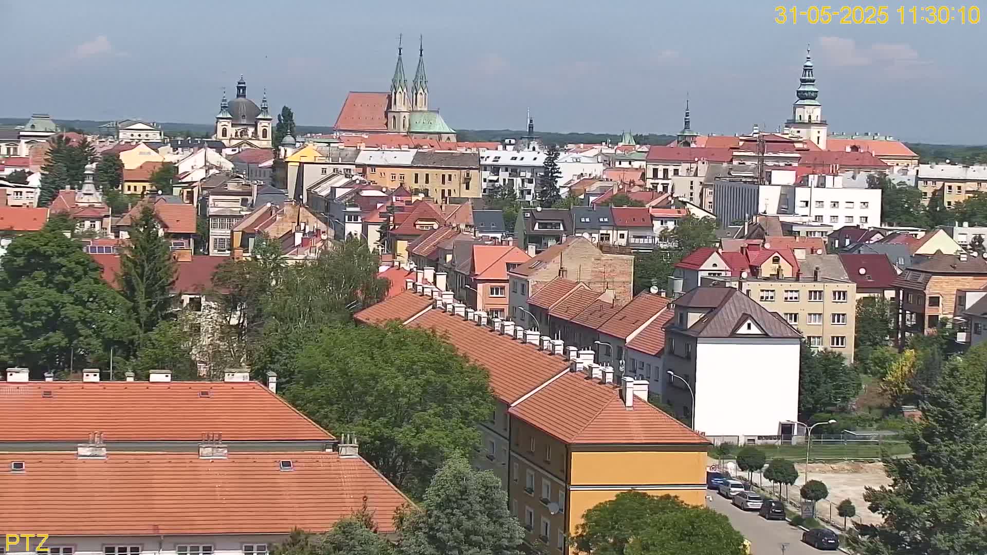 A high-angle, long shot on a sunny day shows a dense collection of red-roofed buildings, interspersed with green trees, in a European city, with several church spires visible in the distance.